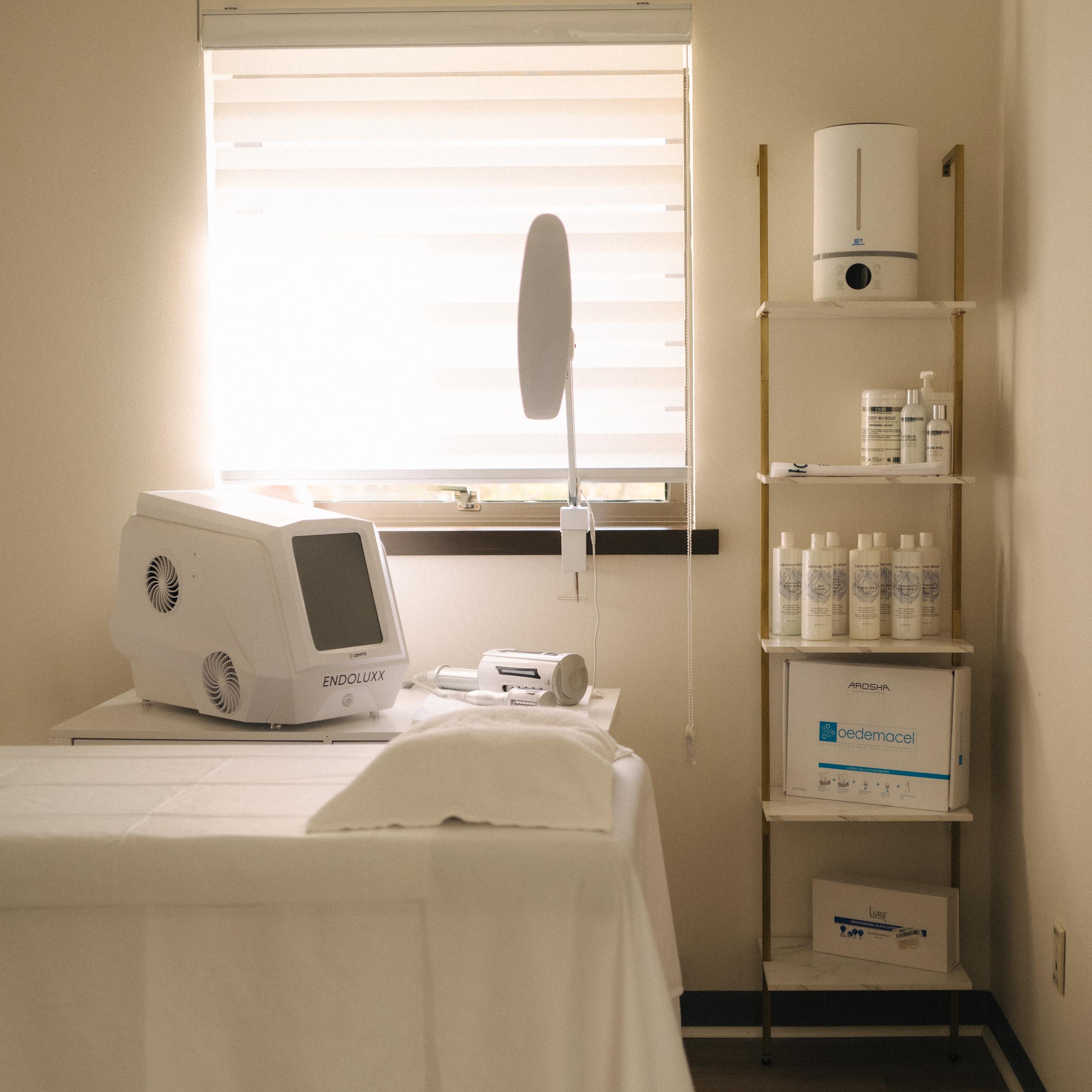 A clean treatment room with a white treatment bed, aesthetic devices, a floor lamp, and shelves stocked with spa products.