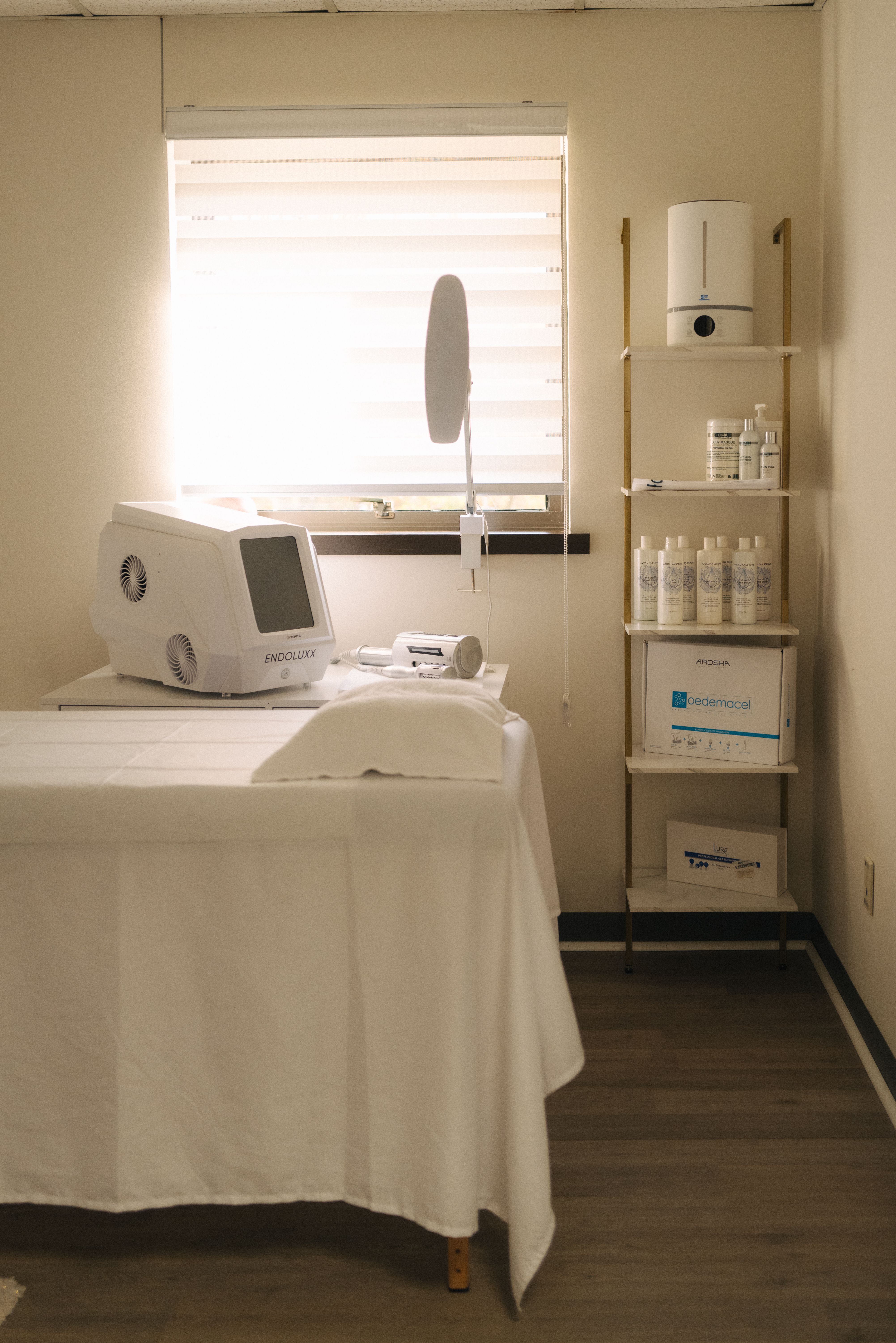 Beauty Republic treatment room with a white treatment bed, facial equipment, and skincare shelves beside a window.