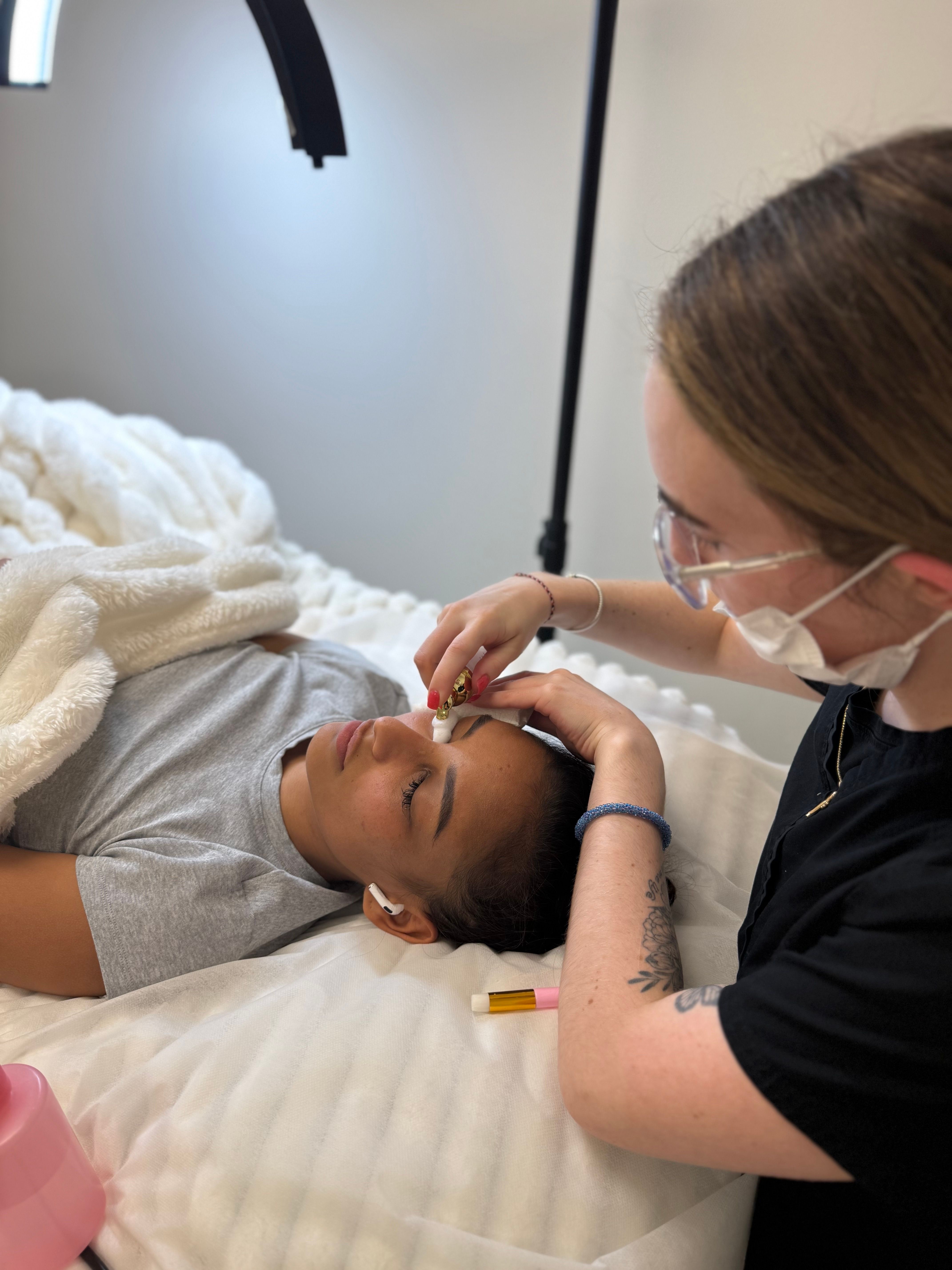 A masked aesthetician washes lashes of a client lying on a bed.