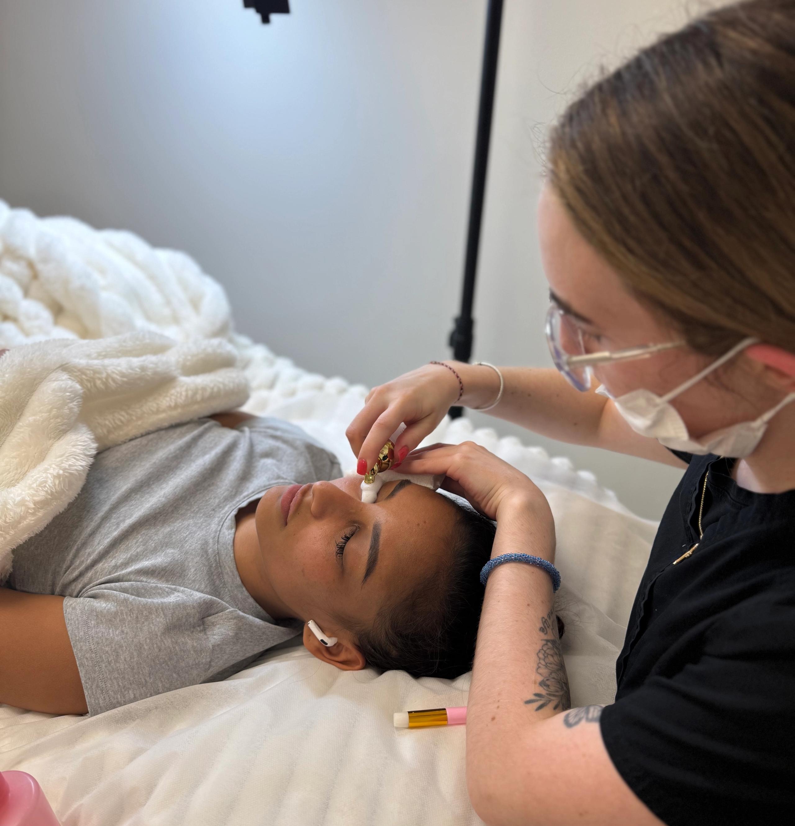 A masked aesthetician washes lashes of a client lying on a bed.