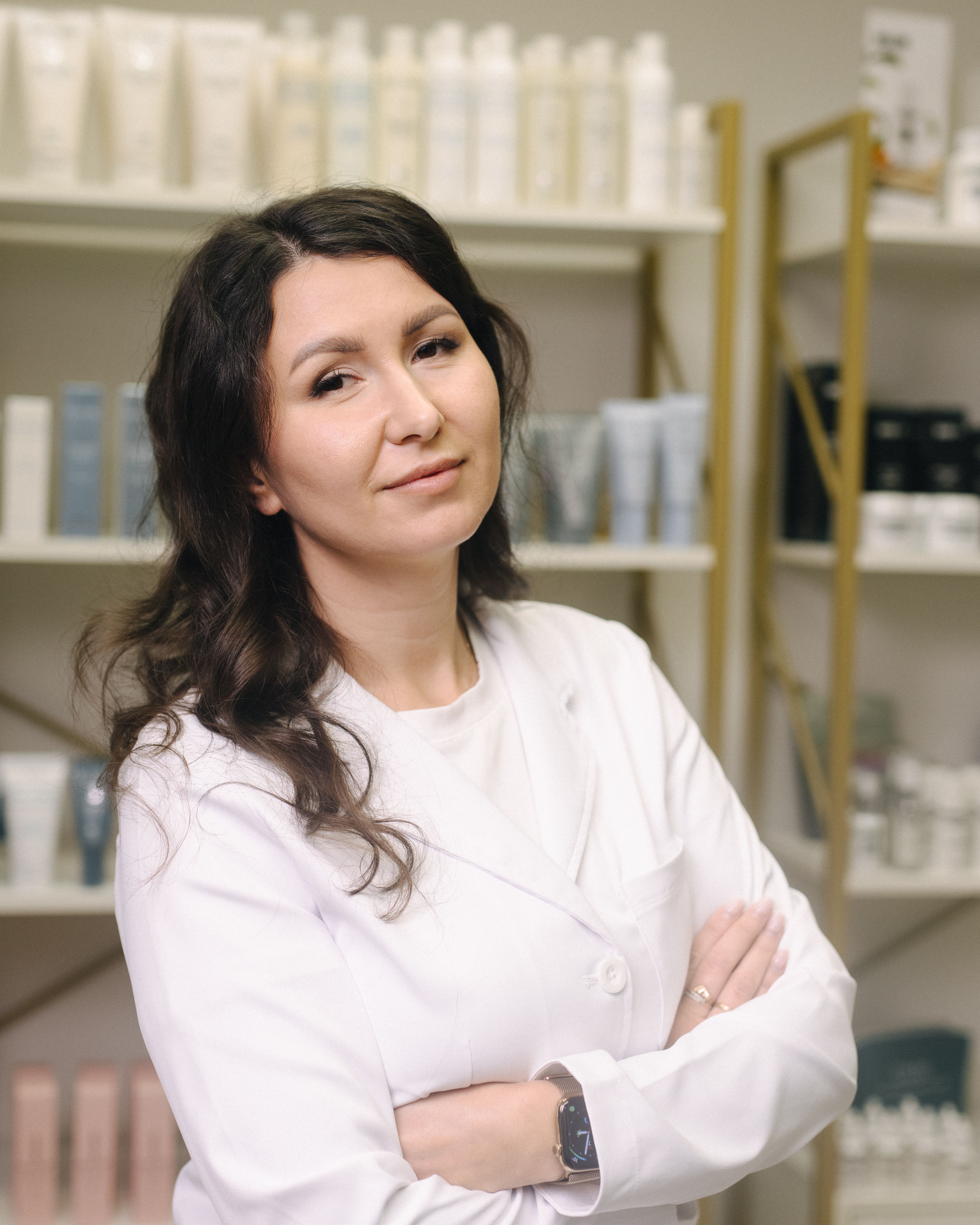 Darya, a Beauty Republic nail artist, in a white coat with her arms crossed beside skincare shelves.