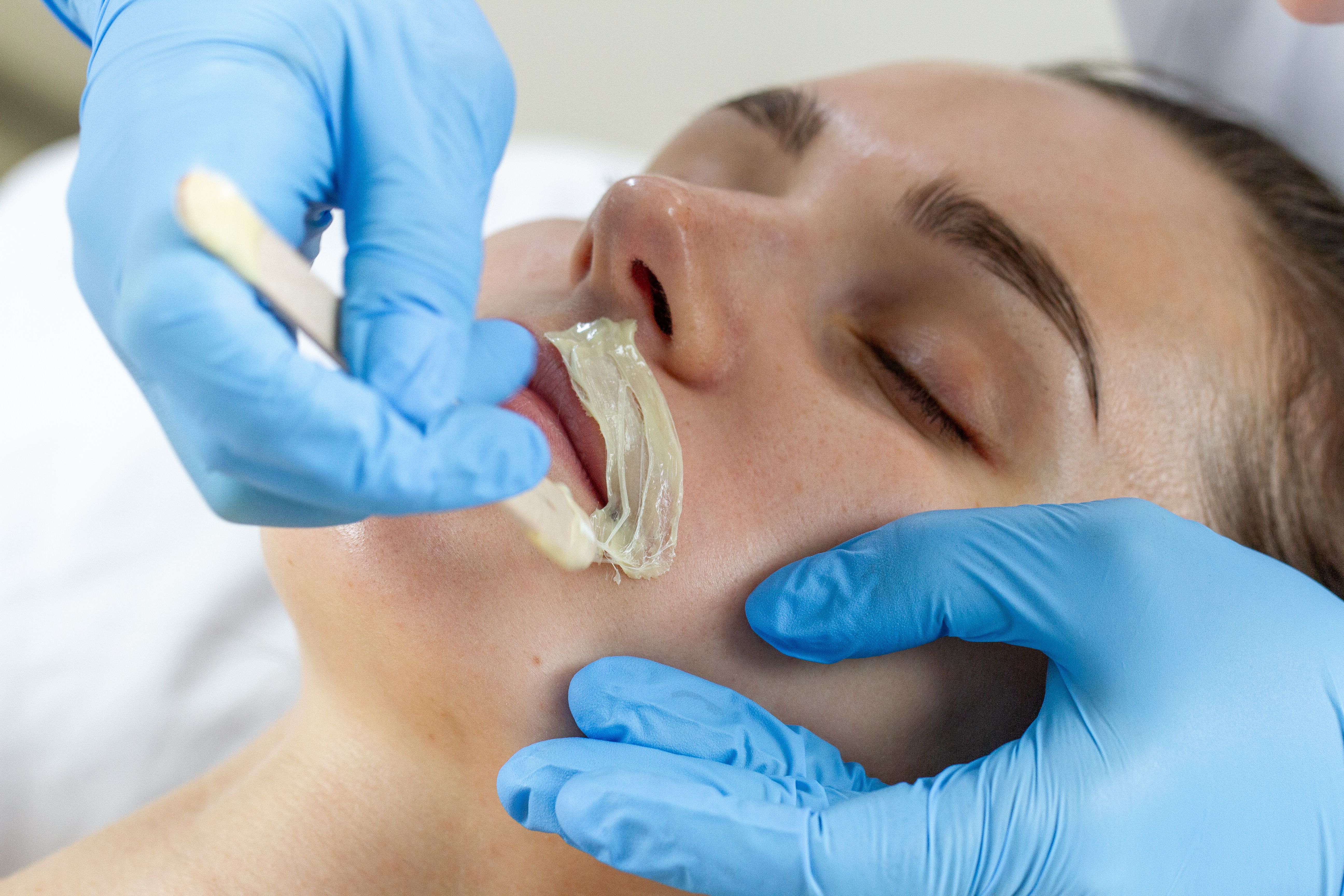 A gloved hand applies wax to a woman's upper lip with an applicator stick.