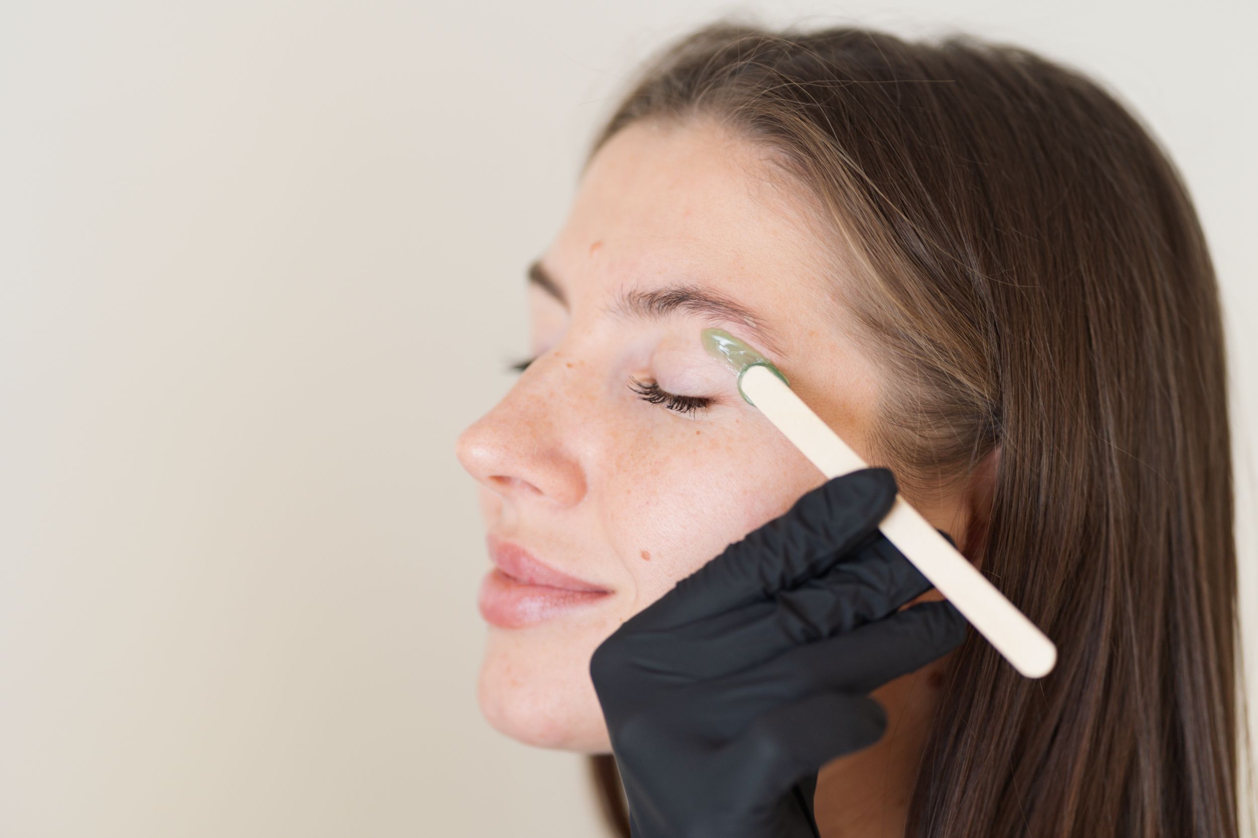 A gloved hand applies wax to a woman's eyebrow with a wooden stick.