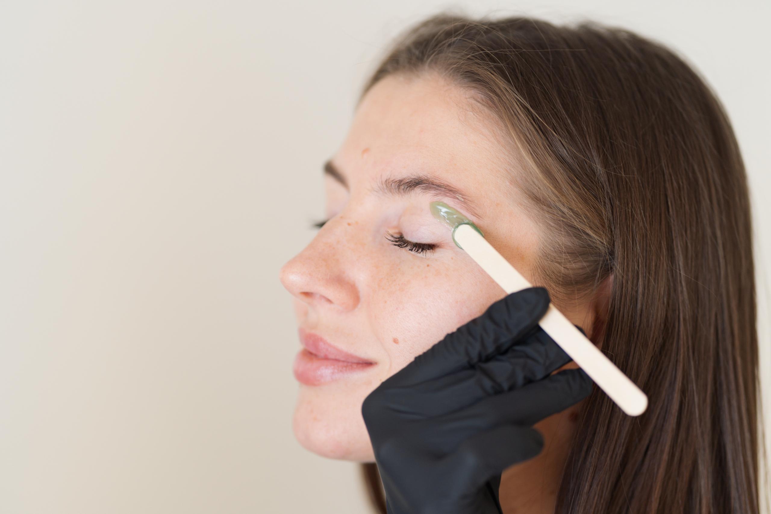 A gloved hand applies wax to a woman's eyebrow with a wooden stick.
