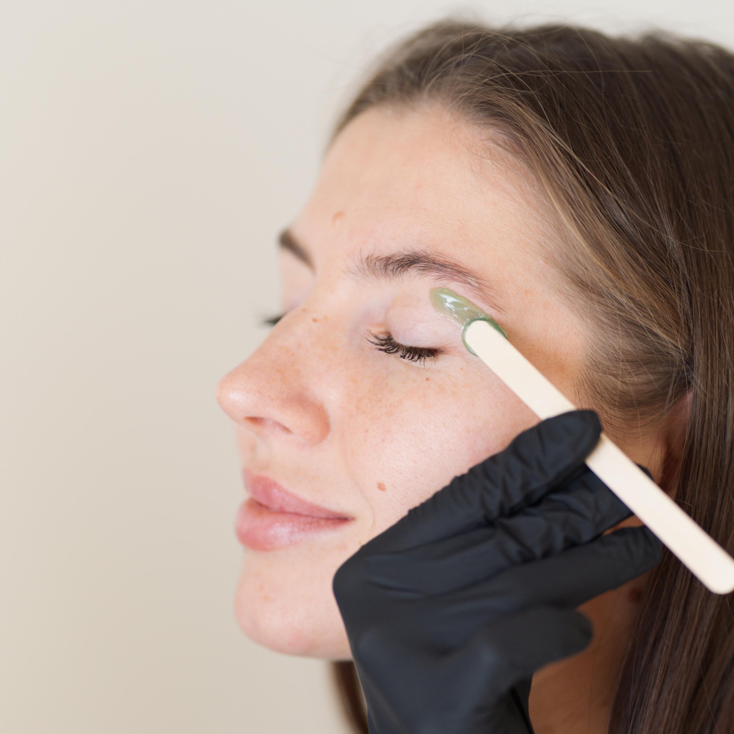 A woman's eyebrow is waxed with green wax applied by a wooden stick.