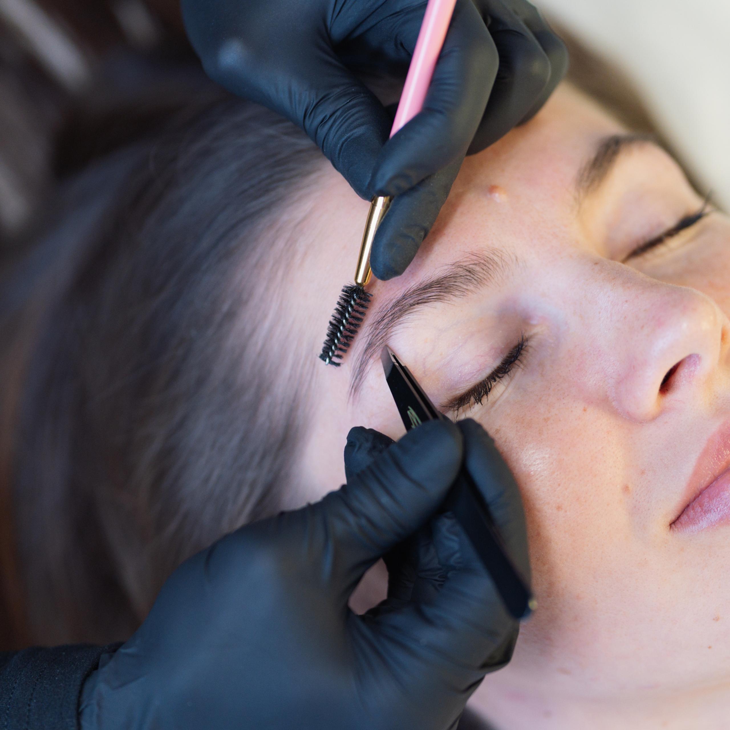 A woman with closed eyes receives an eyebrow grooming treatment from gloved hands.