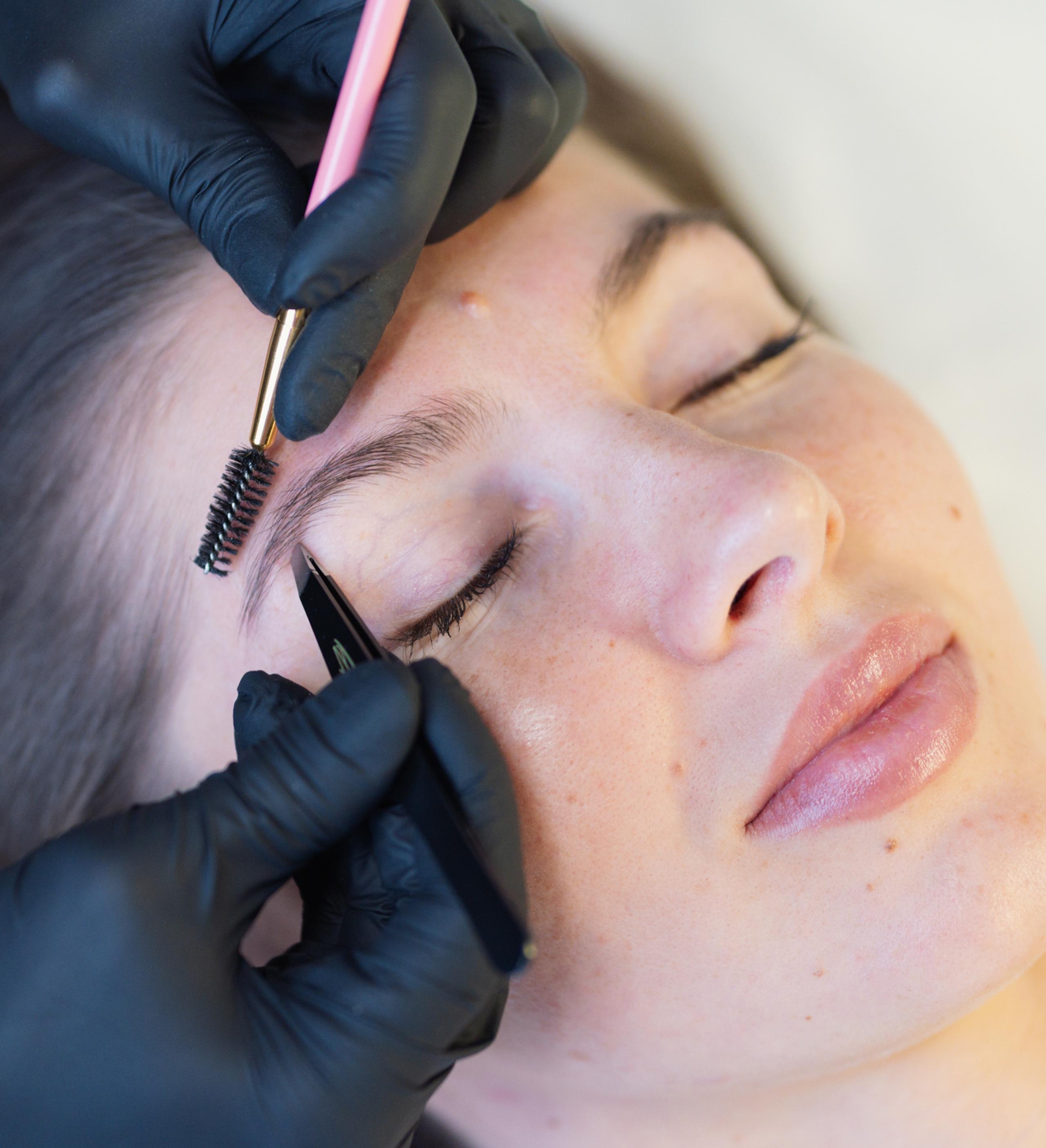 Close-up of a person getting their eyebrows groomed with a spoolie brush and tweezers.