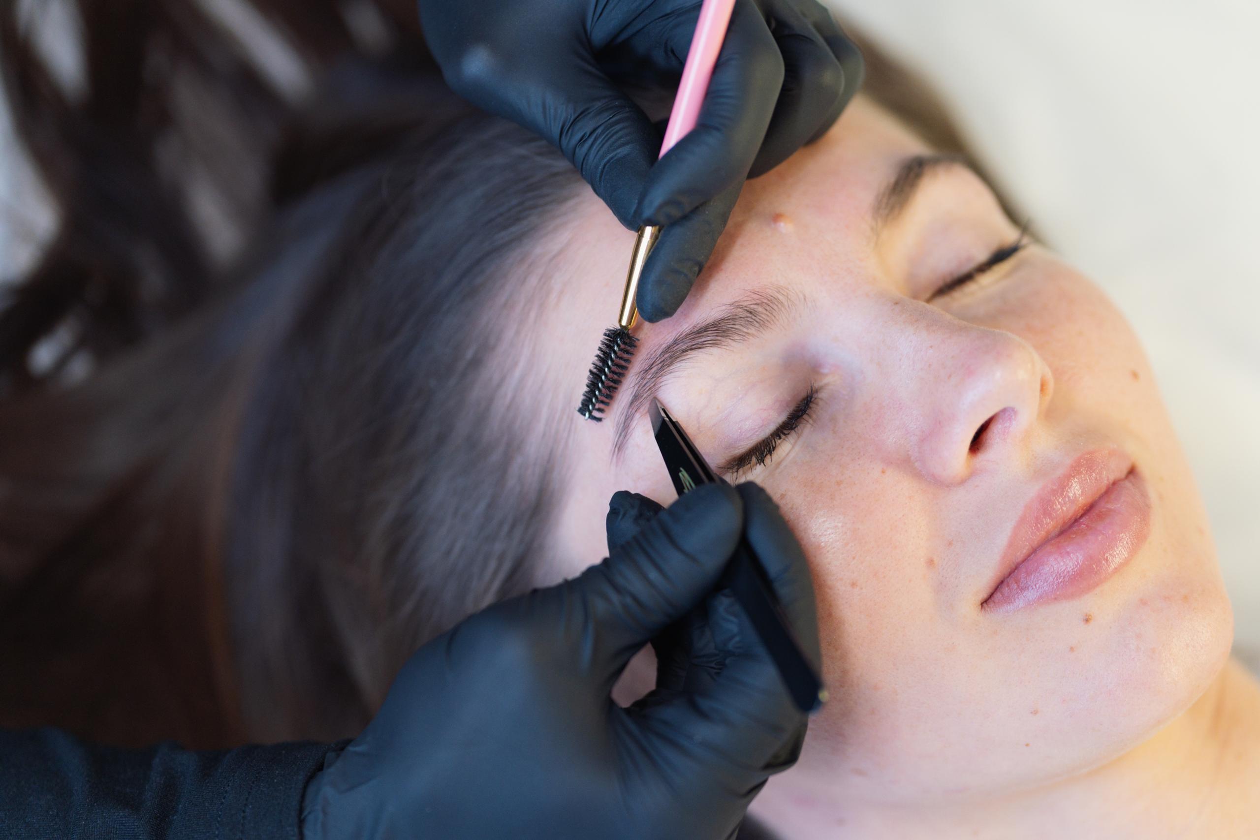 Gloved hands grooming a woman's eyebrows with a brush and tweezers.