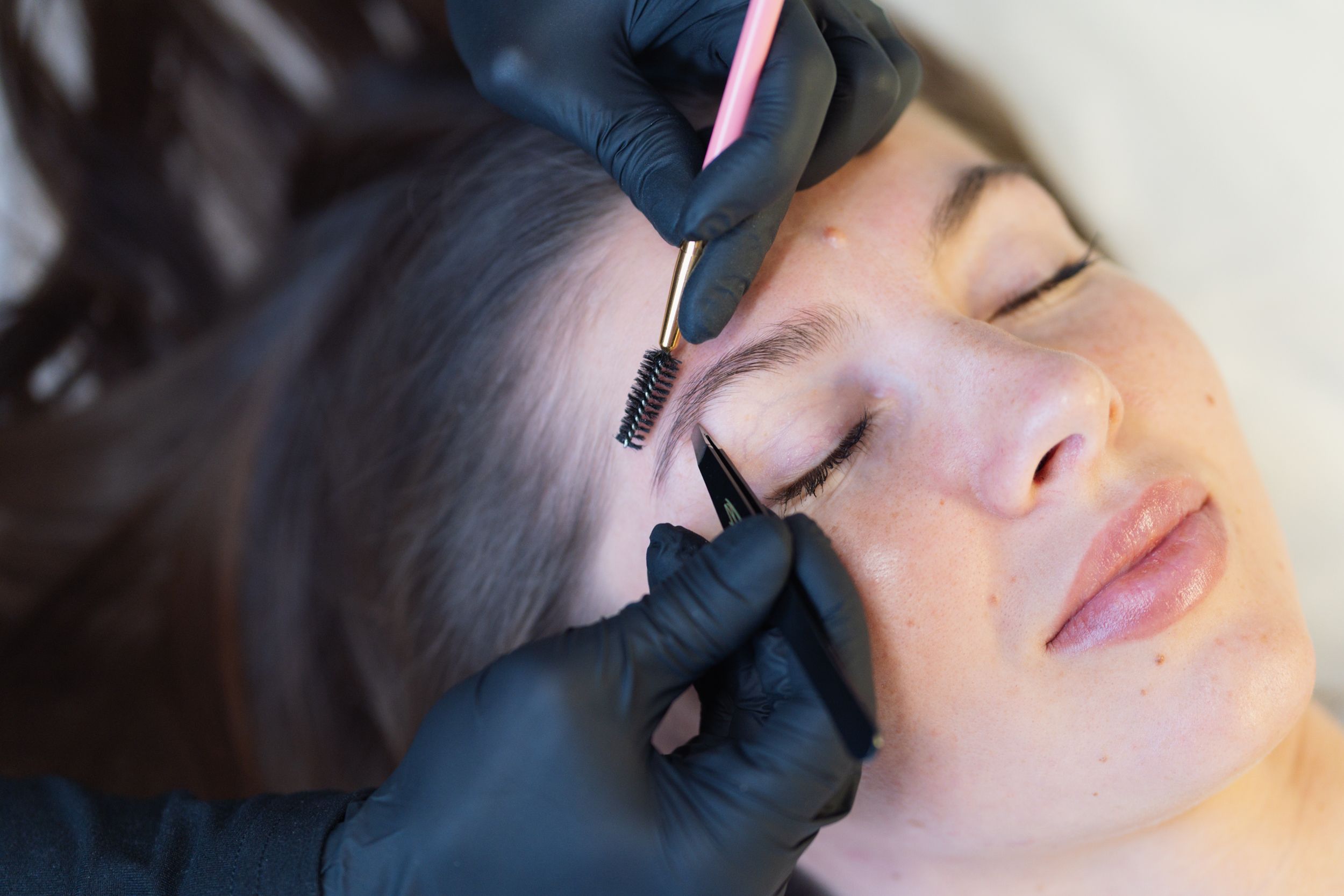 Gloved hands shaping a woman's eyebrows with a brush and tweezers.
