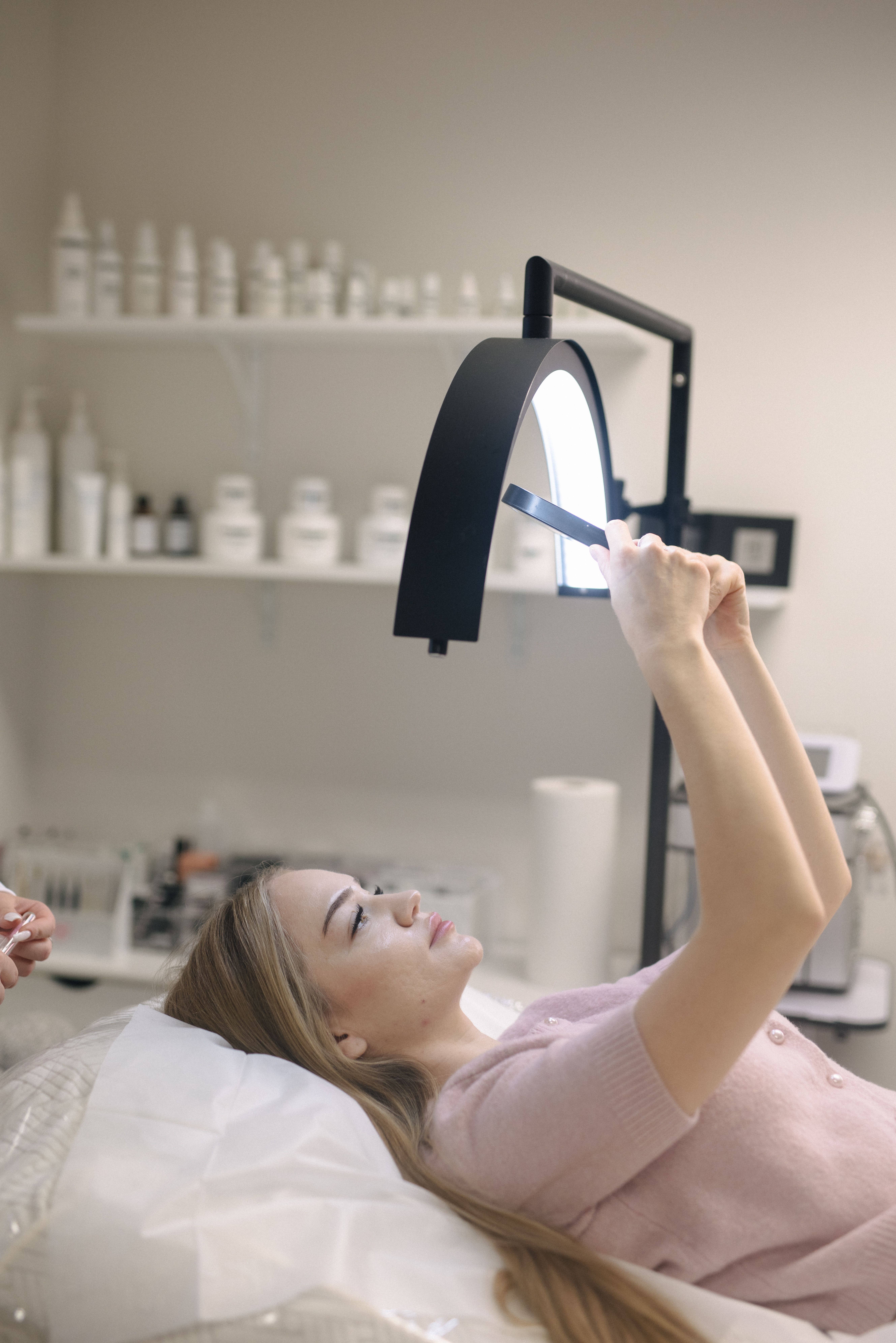 A woman with blonde hair lies on a salon bed, holding up a mirror to view her lashes
