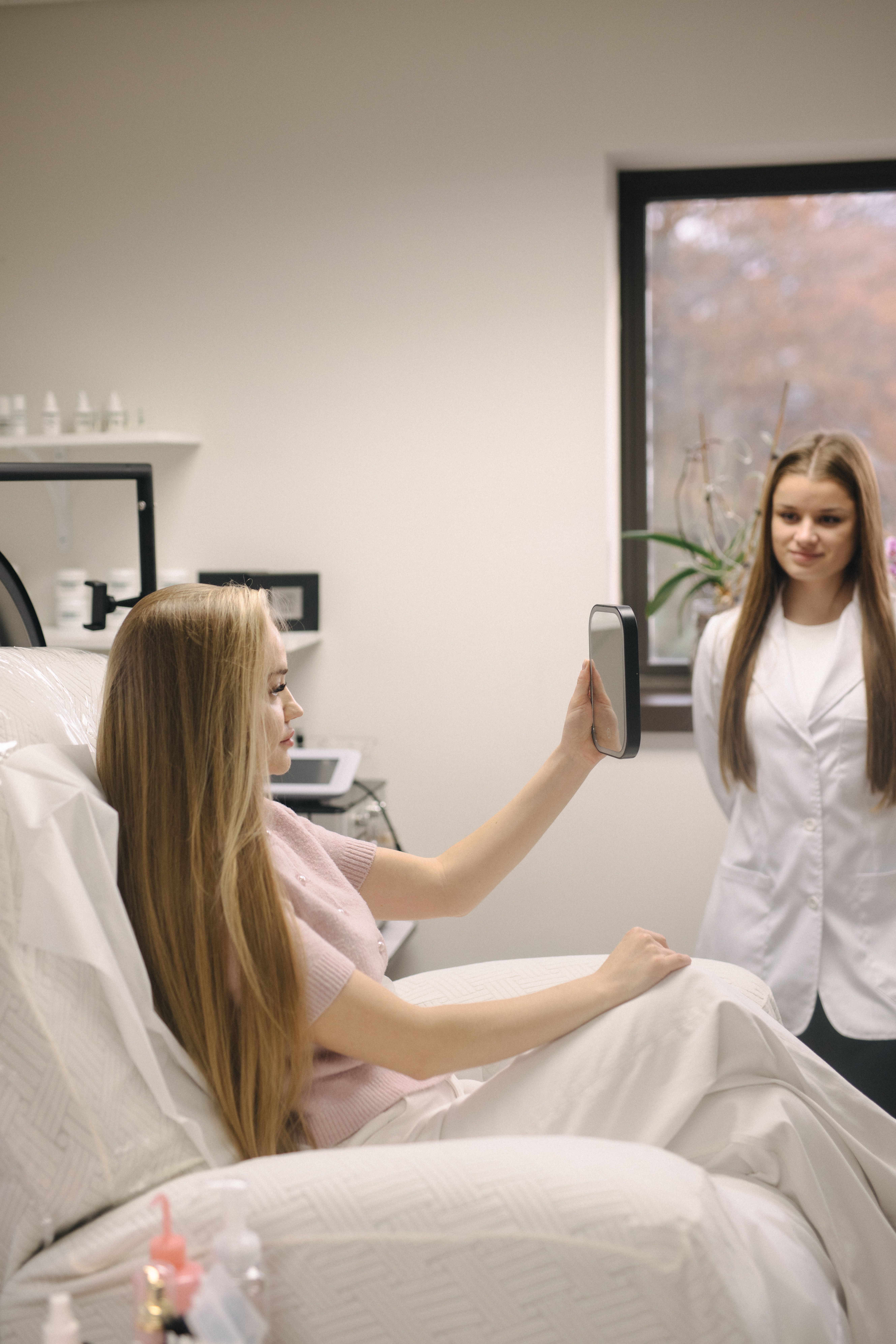 A woman in a treatment chair looks in a mirror while a professional stands nearby.