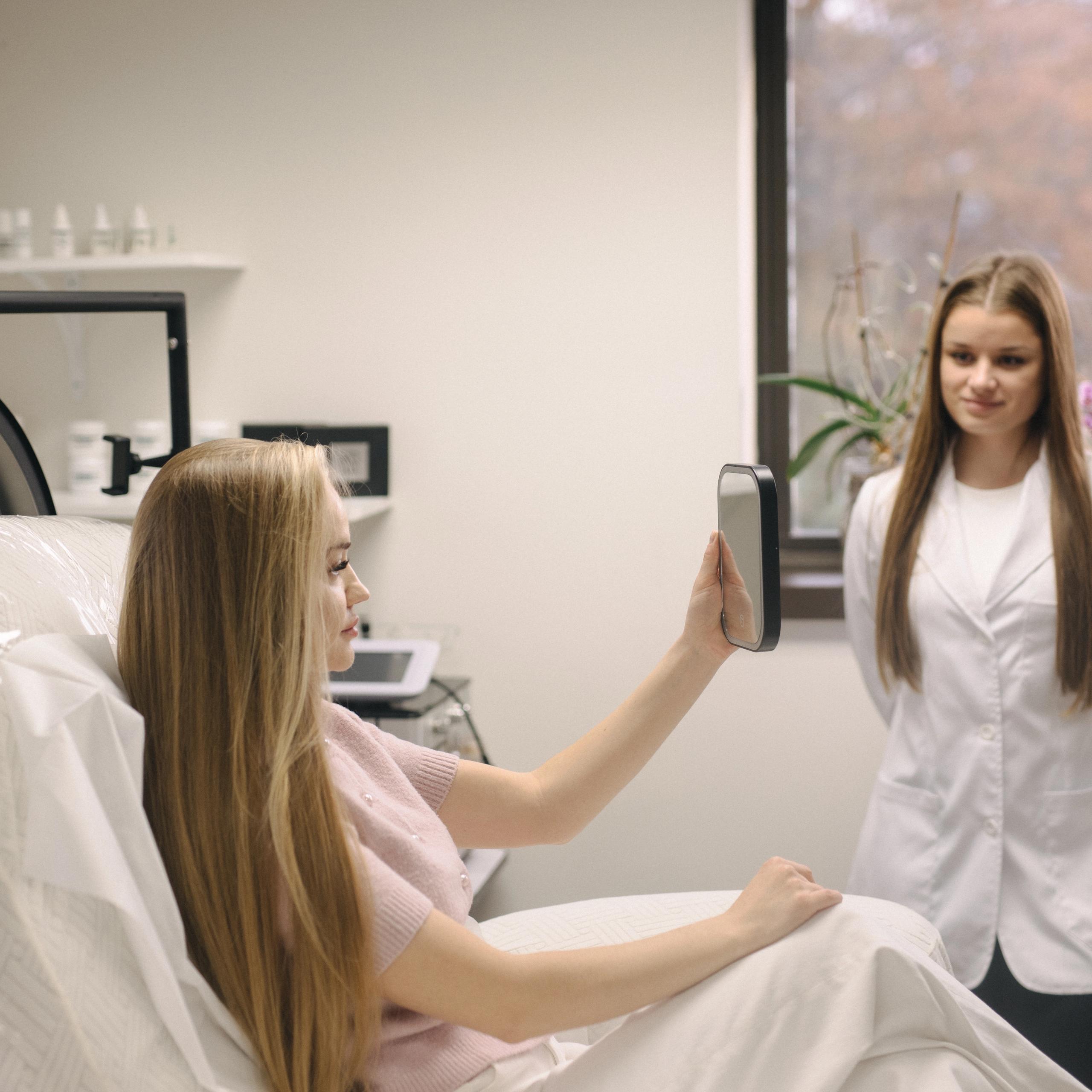 A woman in a treatment chair looks in a mirror while a professional stands nearby.