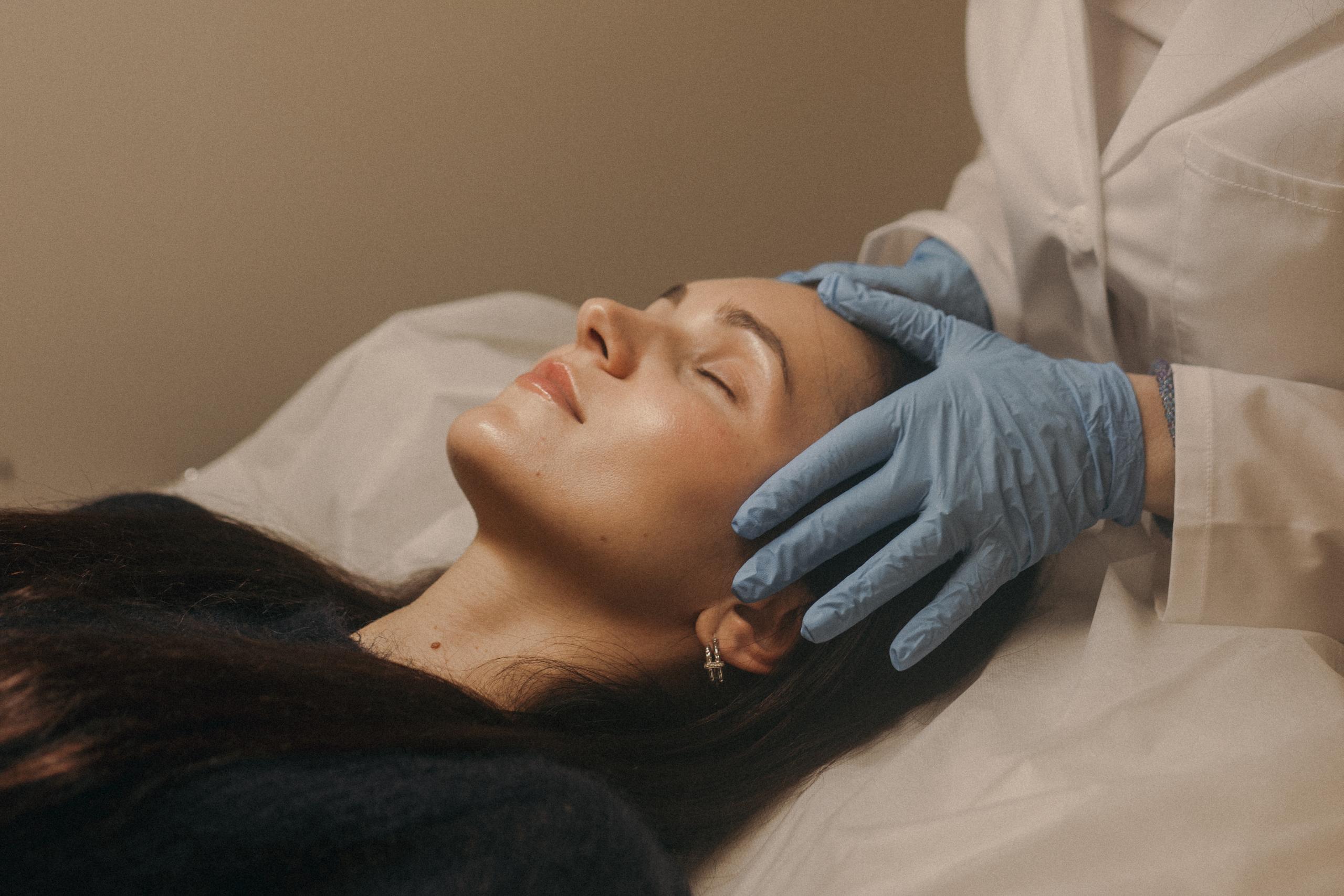 A technician in a lab coat and blue gloves performs a facial treatment on a client lying down under a lamp.
