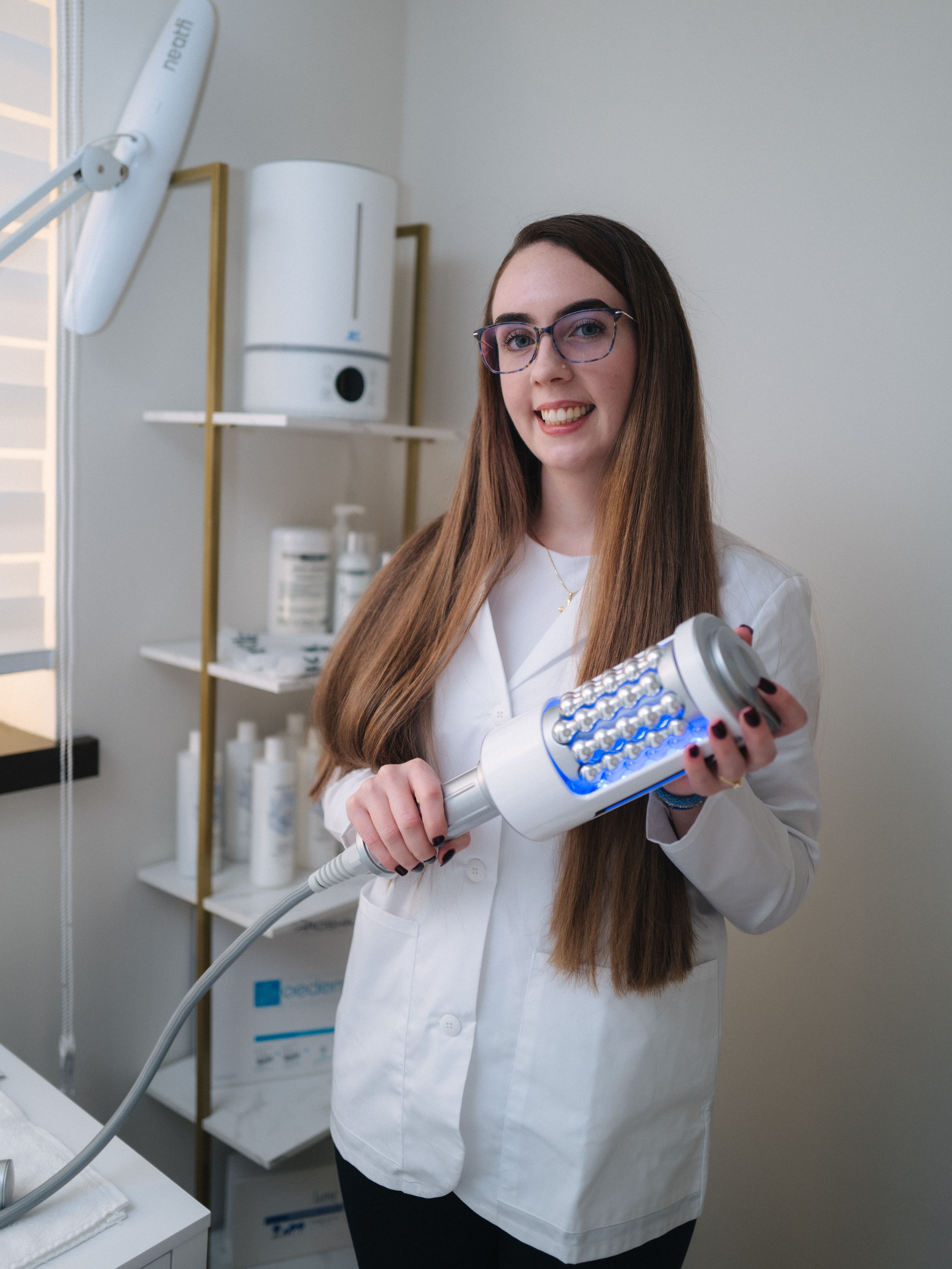 A smiling woman in a lab coat holds a modern beauty device with blue LED lights and rollers.