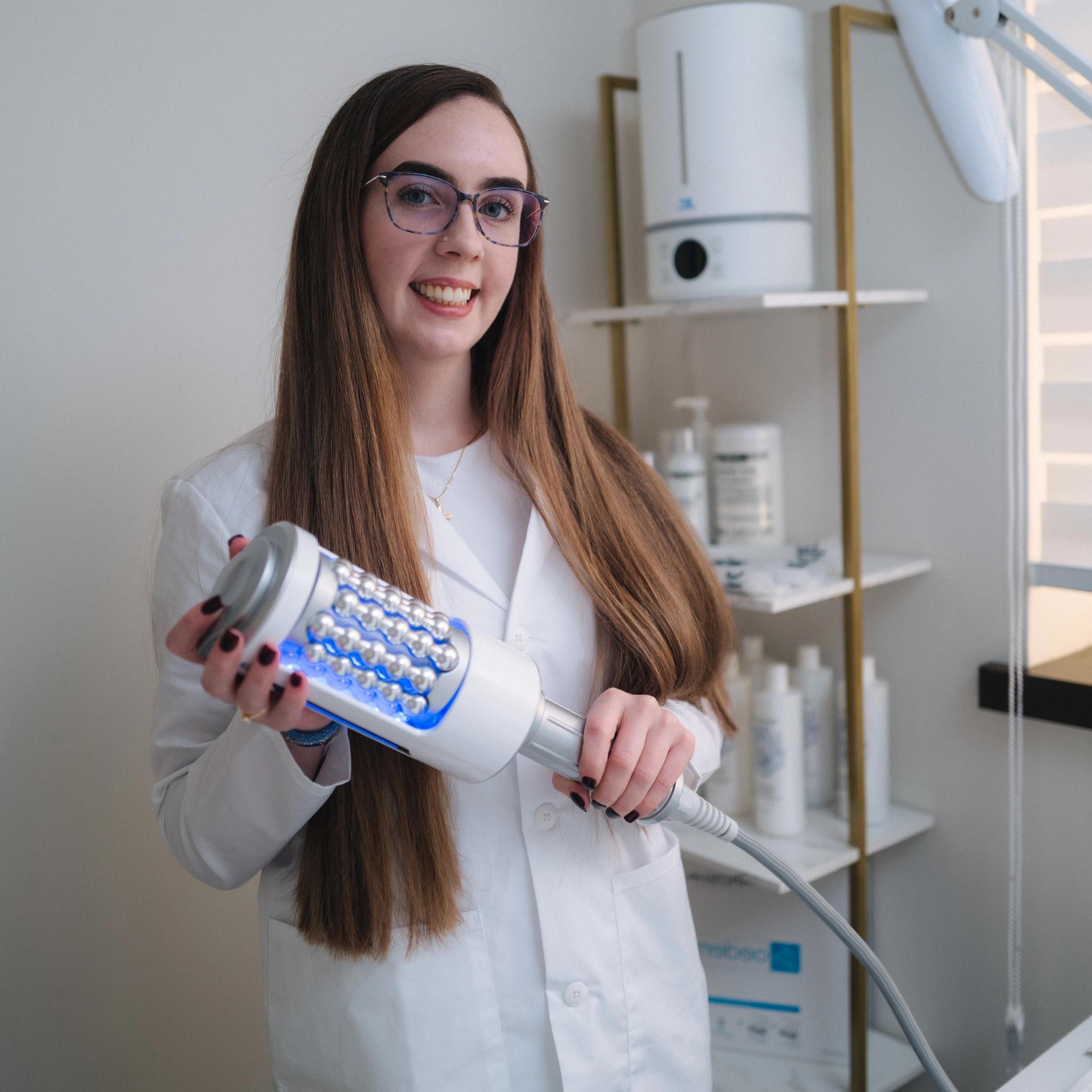 A smiling woman in a lab coat holds a modern beauty device with blue LED lights and rollers.