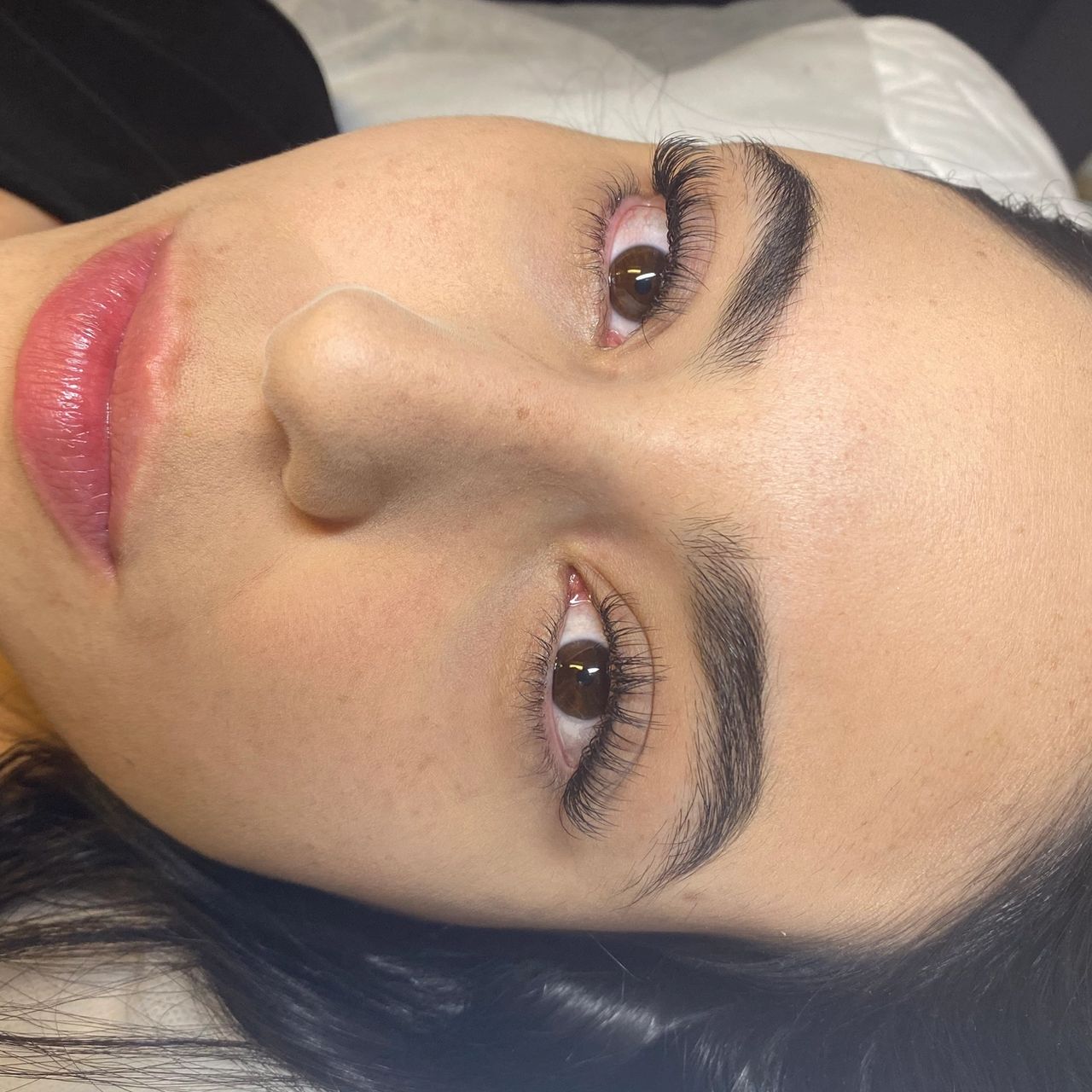 Close-up of a woman lying down with dark brows, long lashes, and pink lipstick.