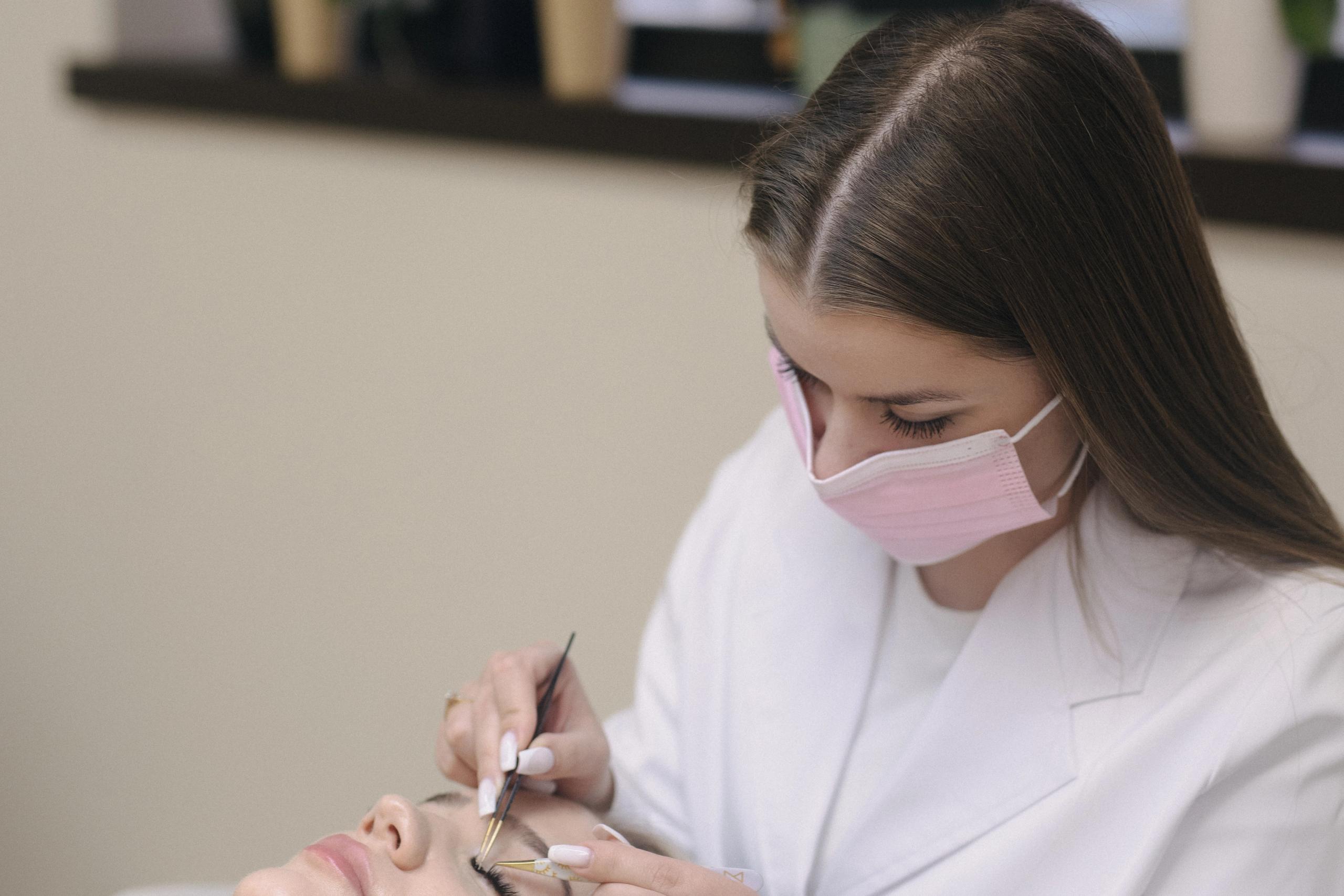 A beautician in a mask applies eyelash extensions to a client.
