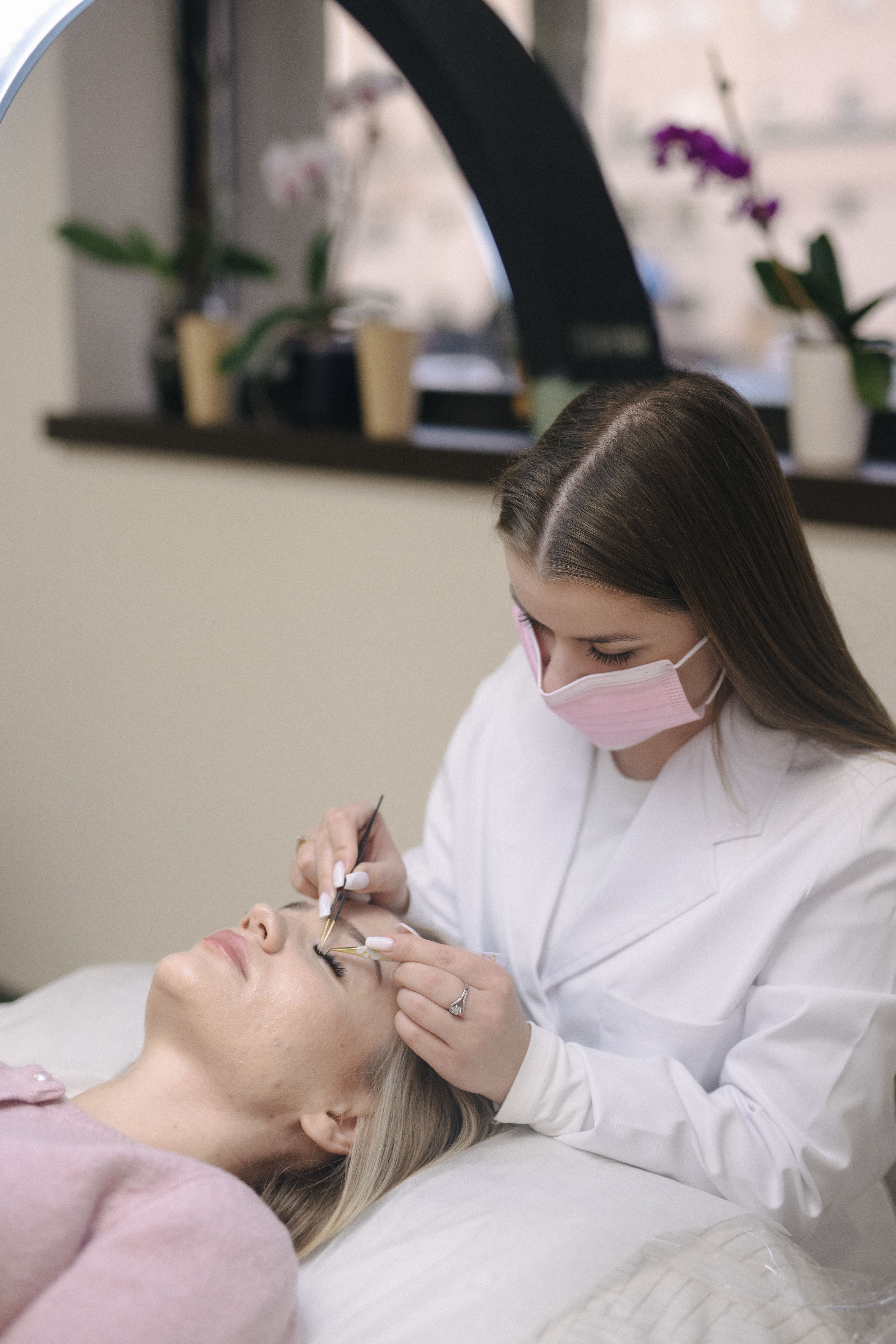 A masked technician in a white coat applies eyelash extensions to a client.