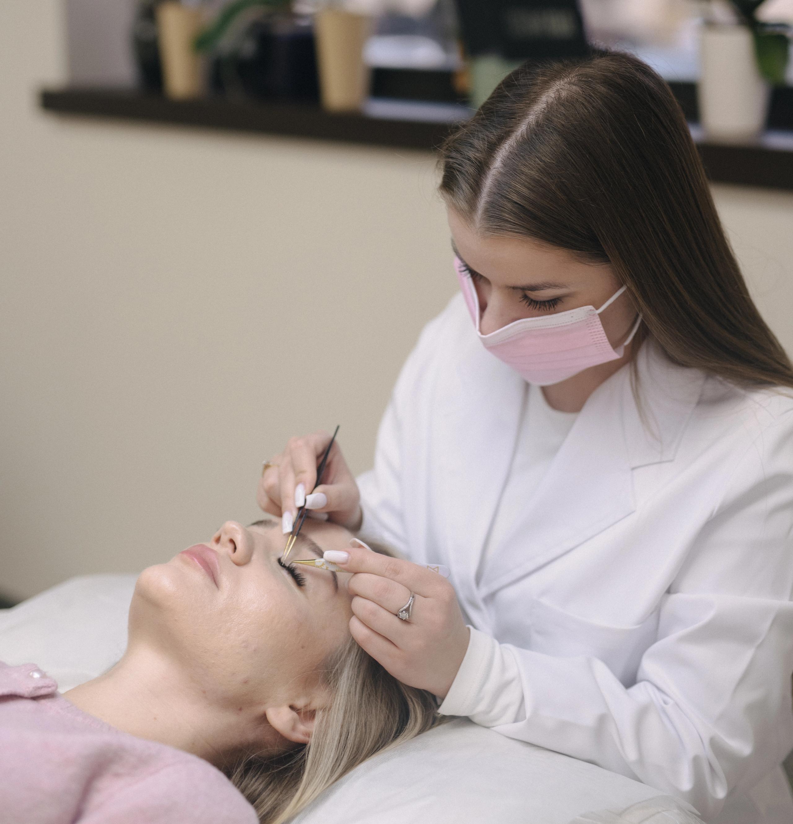 A masked technician in a white coat applies eyelash extensions to a client.
