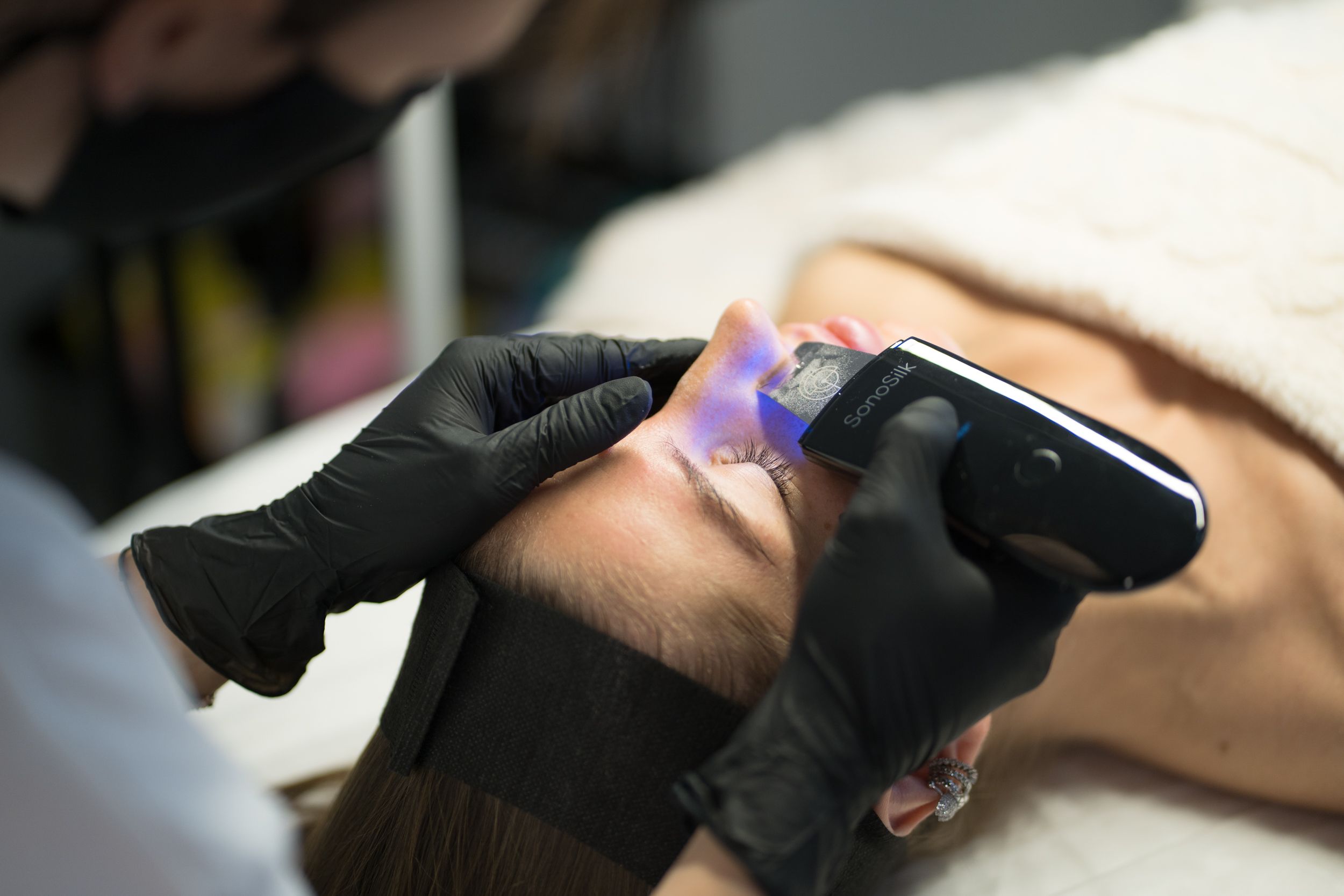 A person receives a facial treatment with a SonaSilk device emitting blue light.