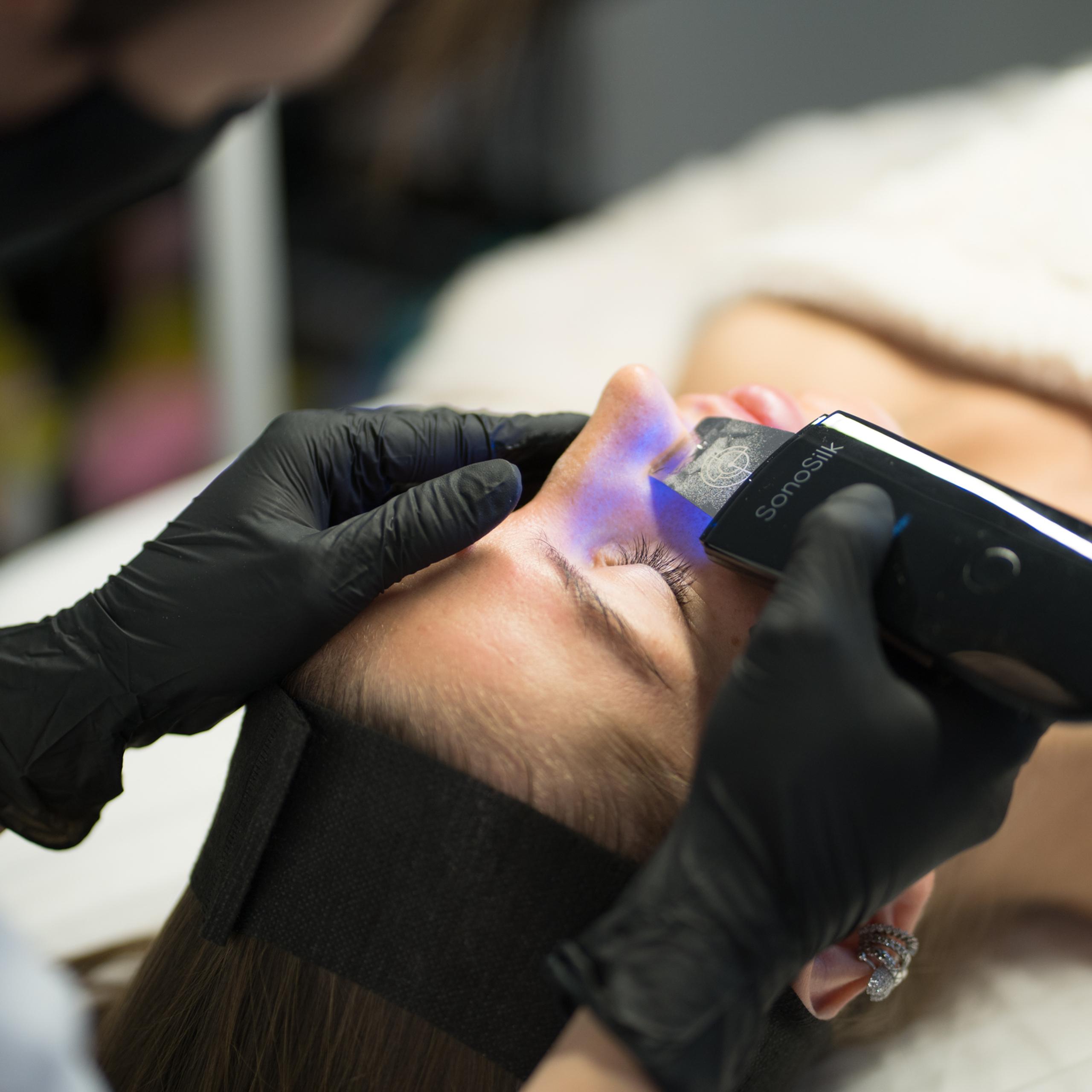A person receives a facial treatment with a SonaSilk device emitting blue light.