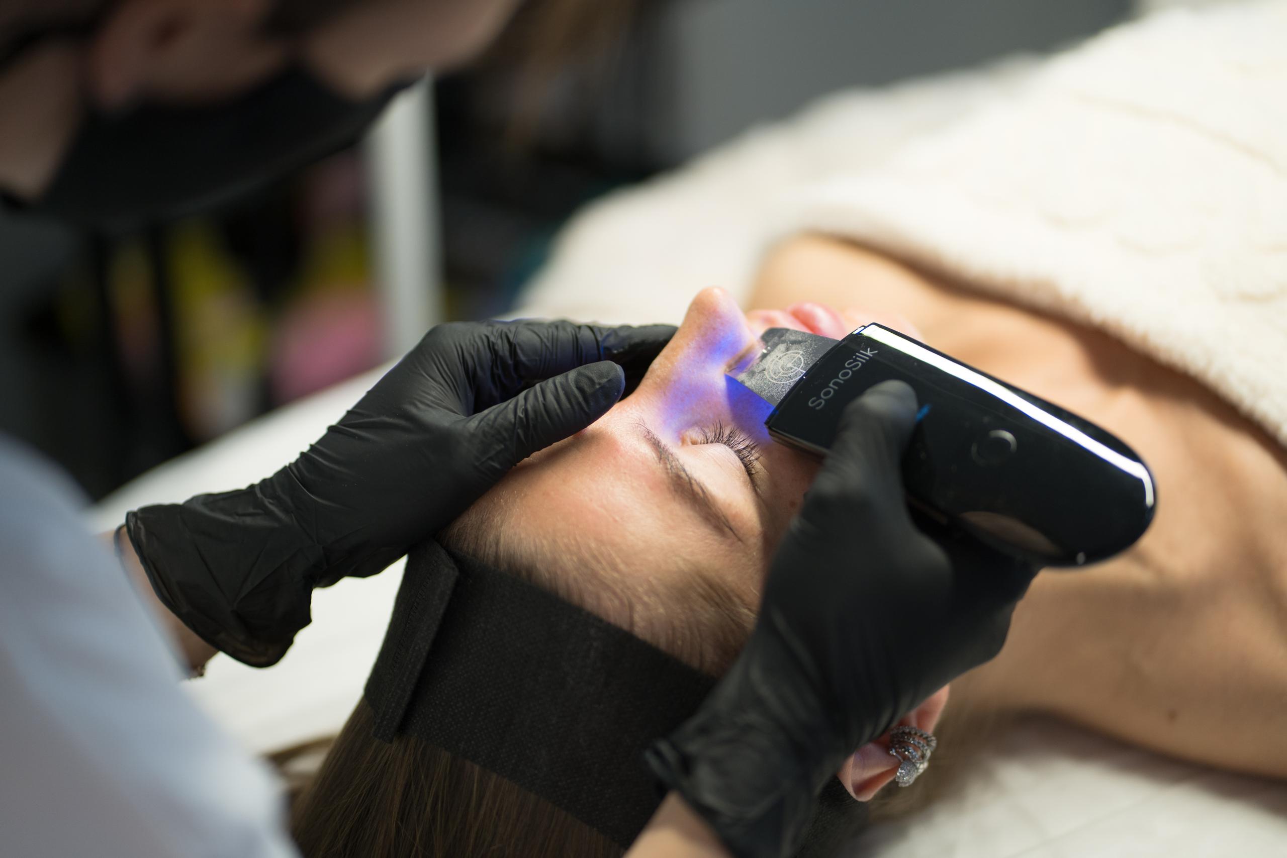 A professional uses a SonaSilk ultrasonic skin spatula with blue light on a client's face.