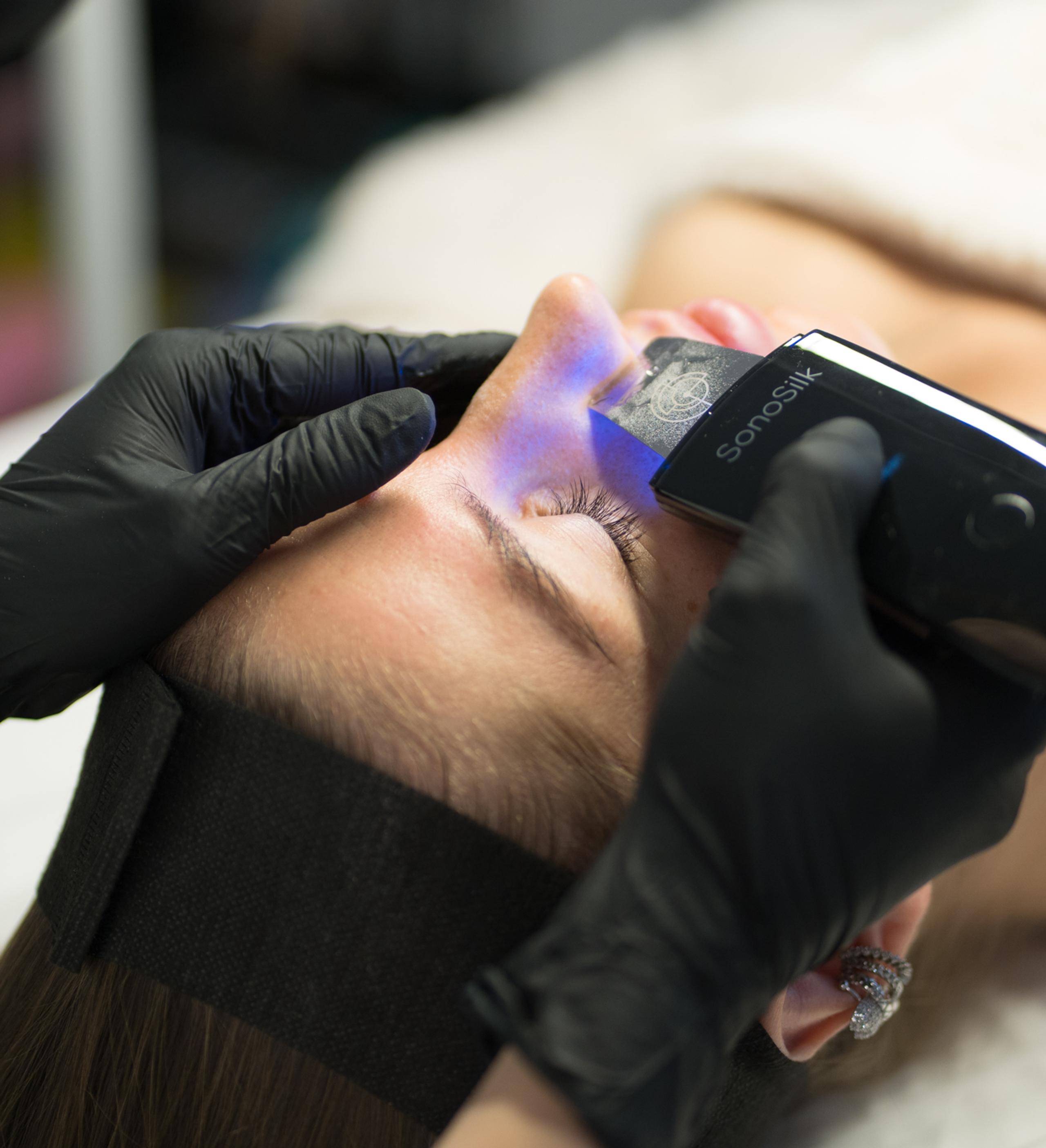 Gloved esthetician gliding a handheld skin device over a client’s forehead during a facial.