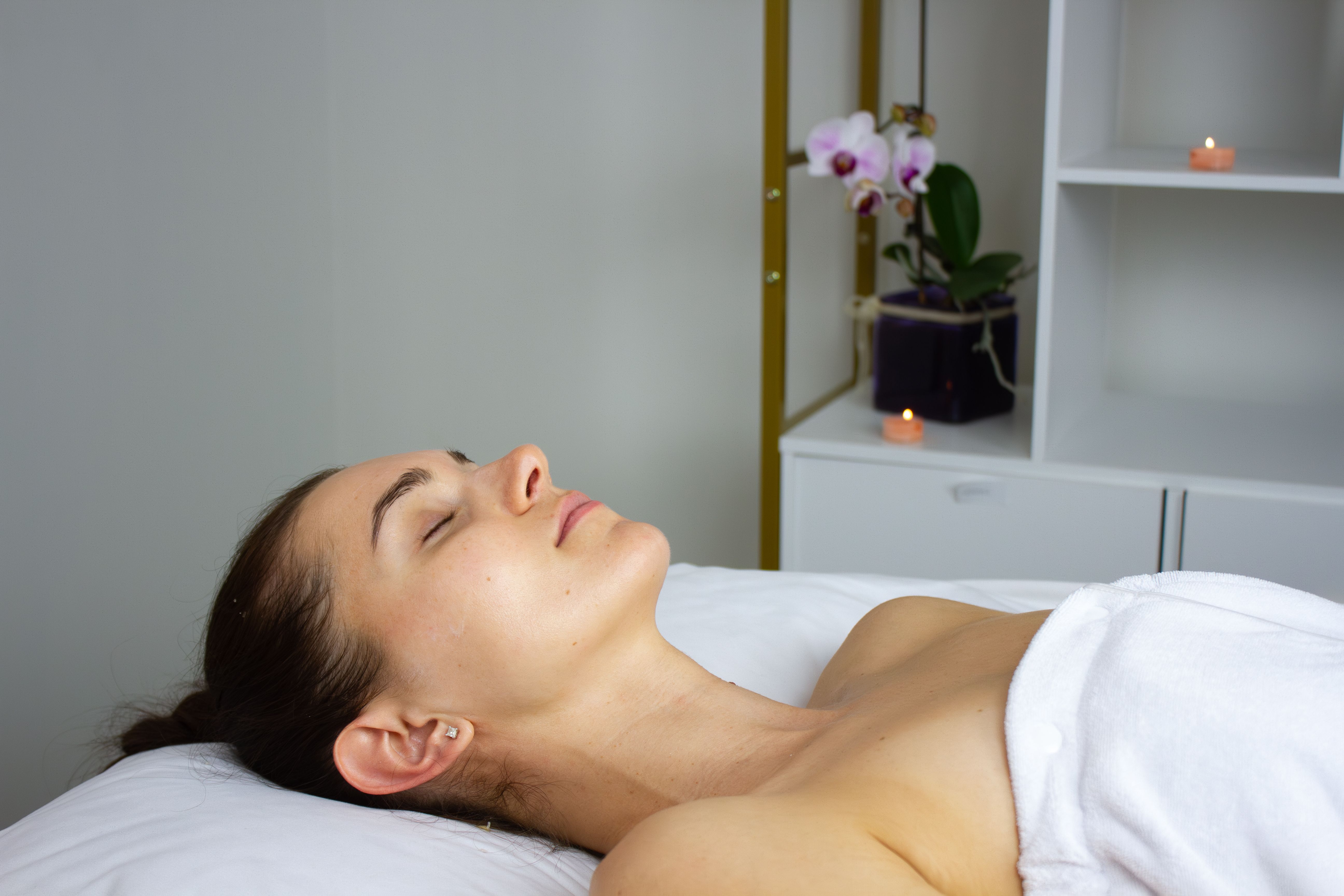 A woman lies peacefully on a spa bed with eyes closed, an orchid and lit candles in the background.