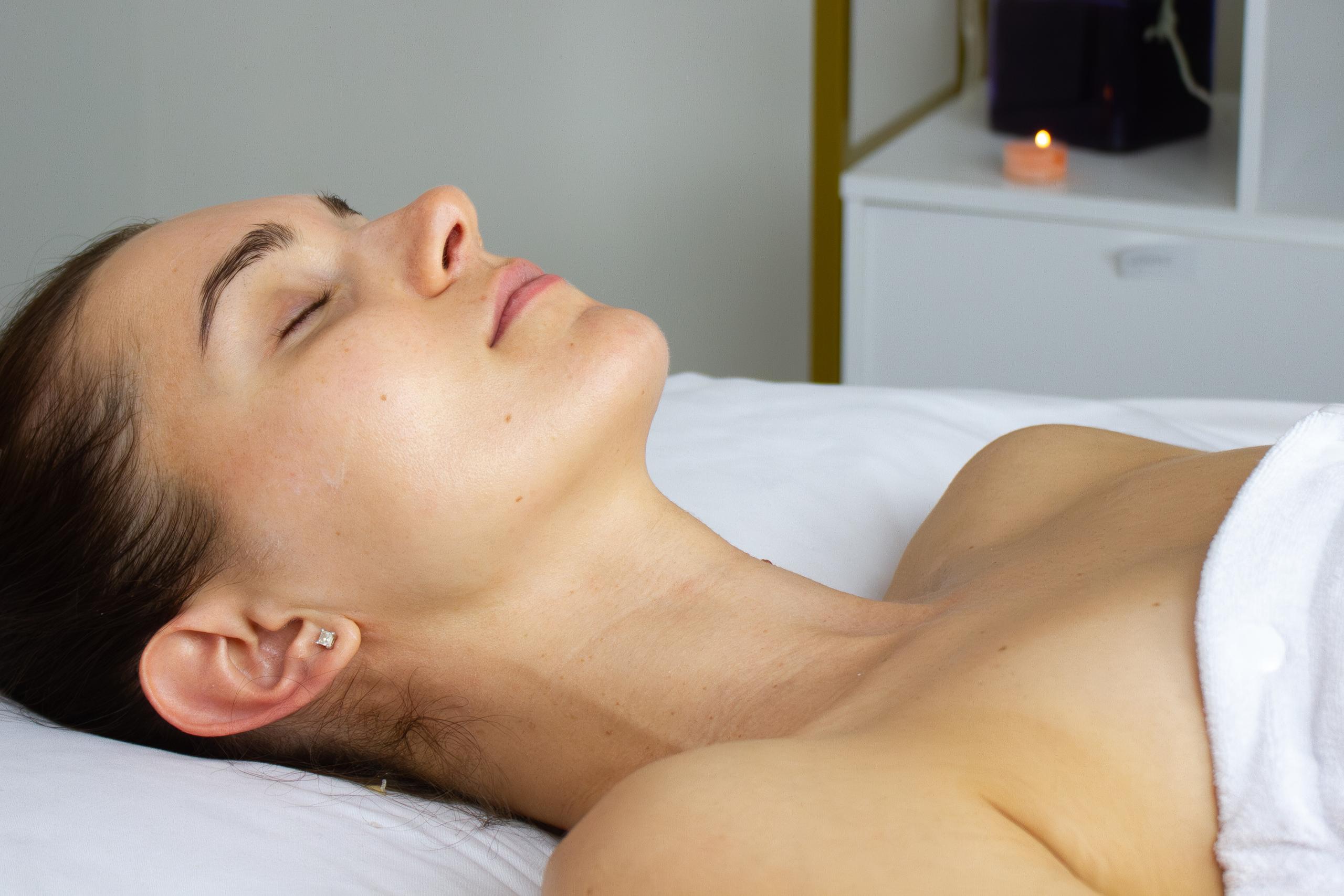A woman lies peacefully on a spa bed with eyes closed, an orchid and lit candles in the background.