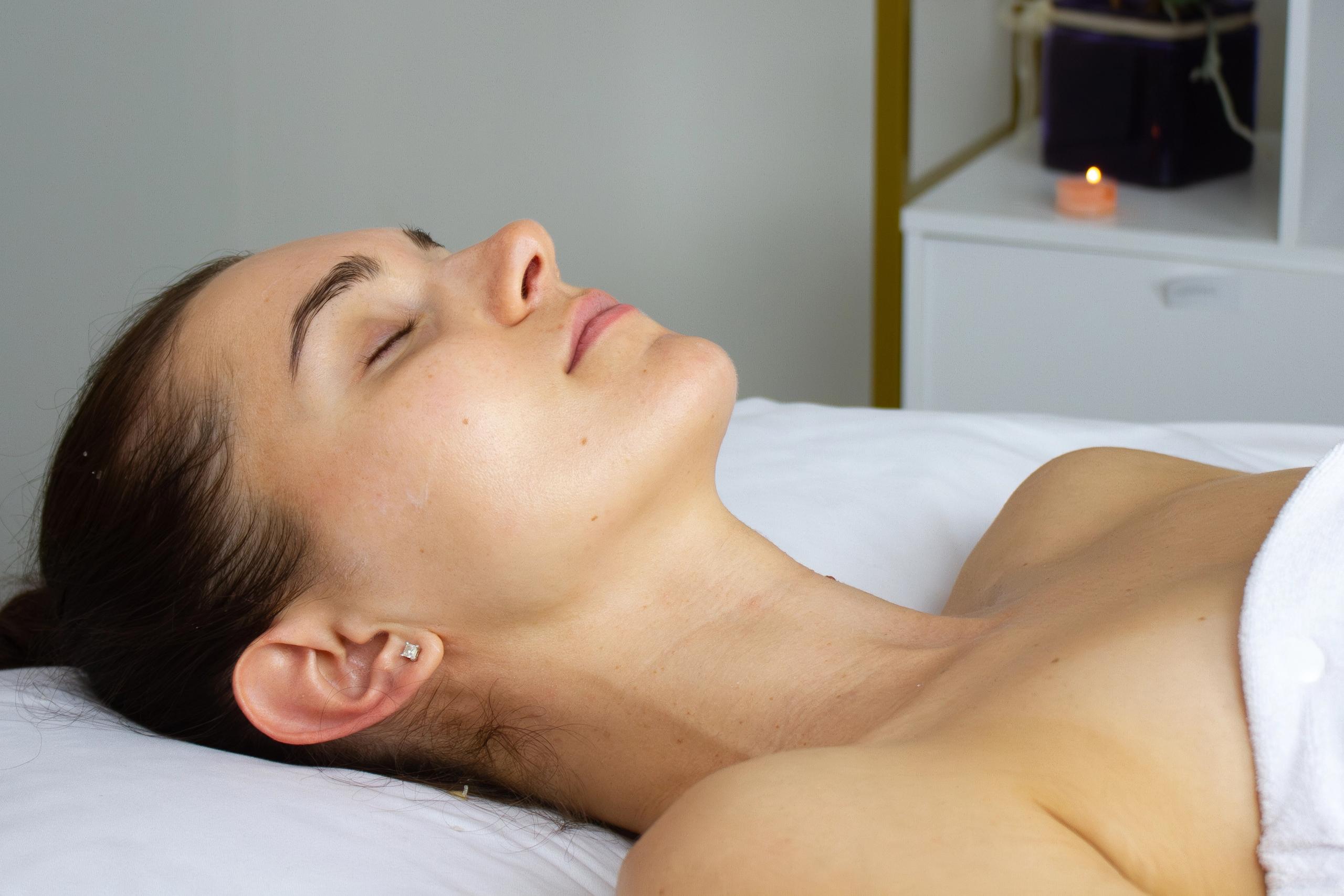 A woman with a soft, relaxed smile and closed eyes is laying on the esthetic bed in a spa setting