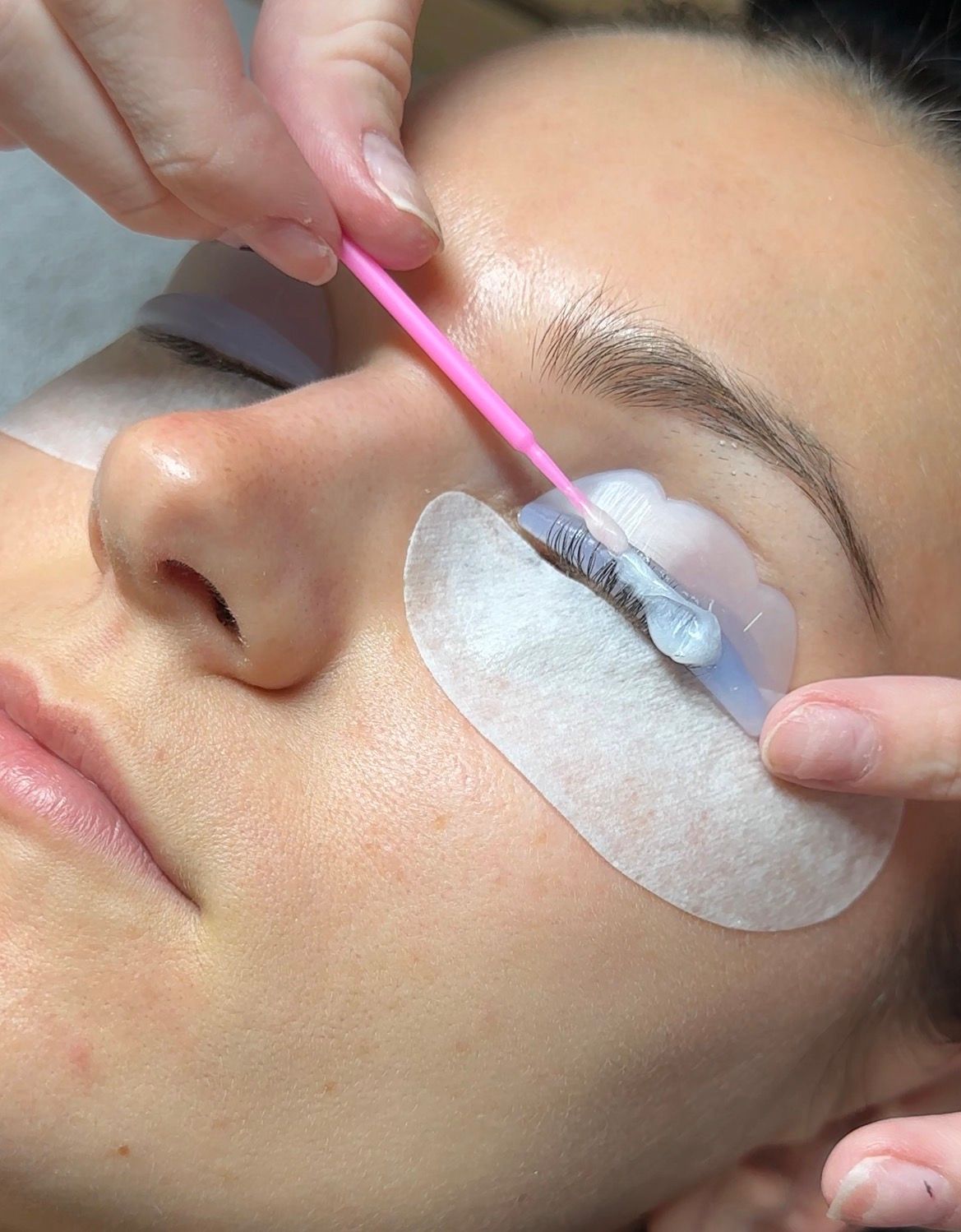 Close-up of a person receiving an eyelash lift, with a technician applying a white cream to their curled lashes using a pink micro-brush.