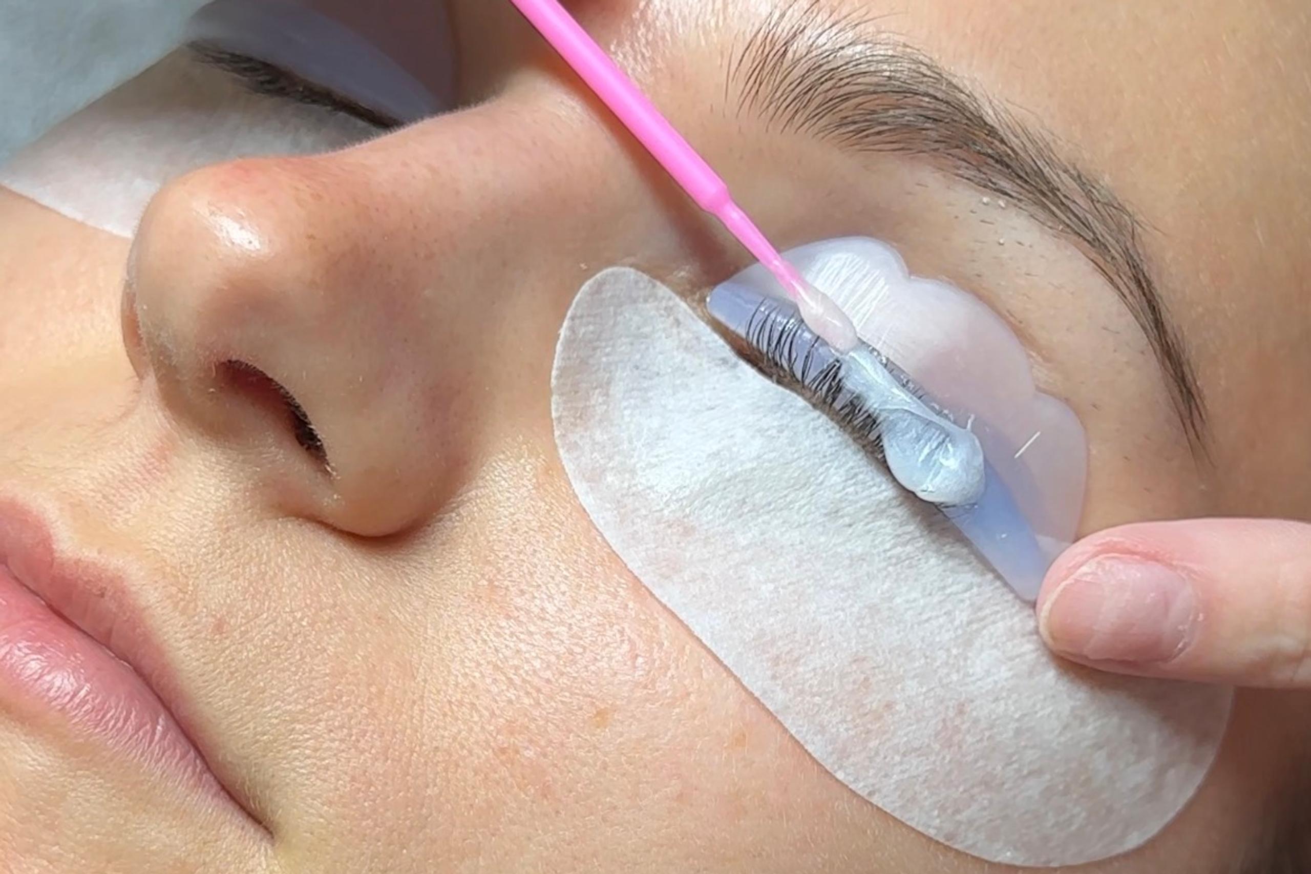 Close-up of a person receiving an eyelash lift, with a technician applying a white cream to their curled lashes using a pink micro-brush.