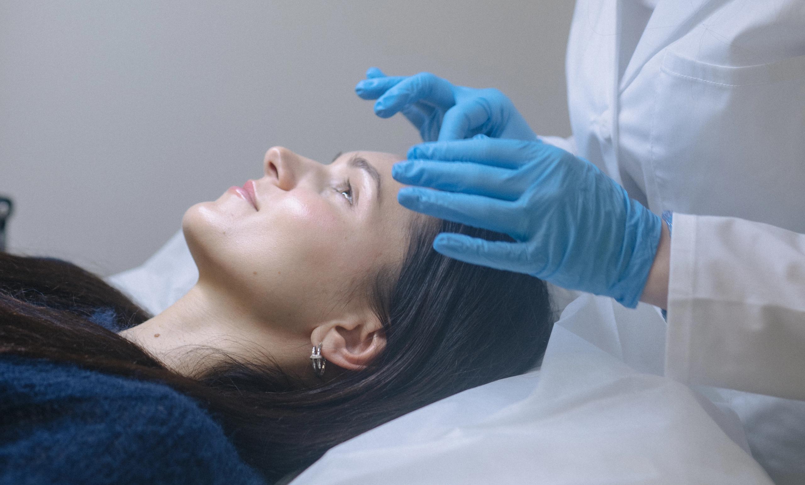 A person in blue gloves and a white lab coat gently touches a woman's forehead.