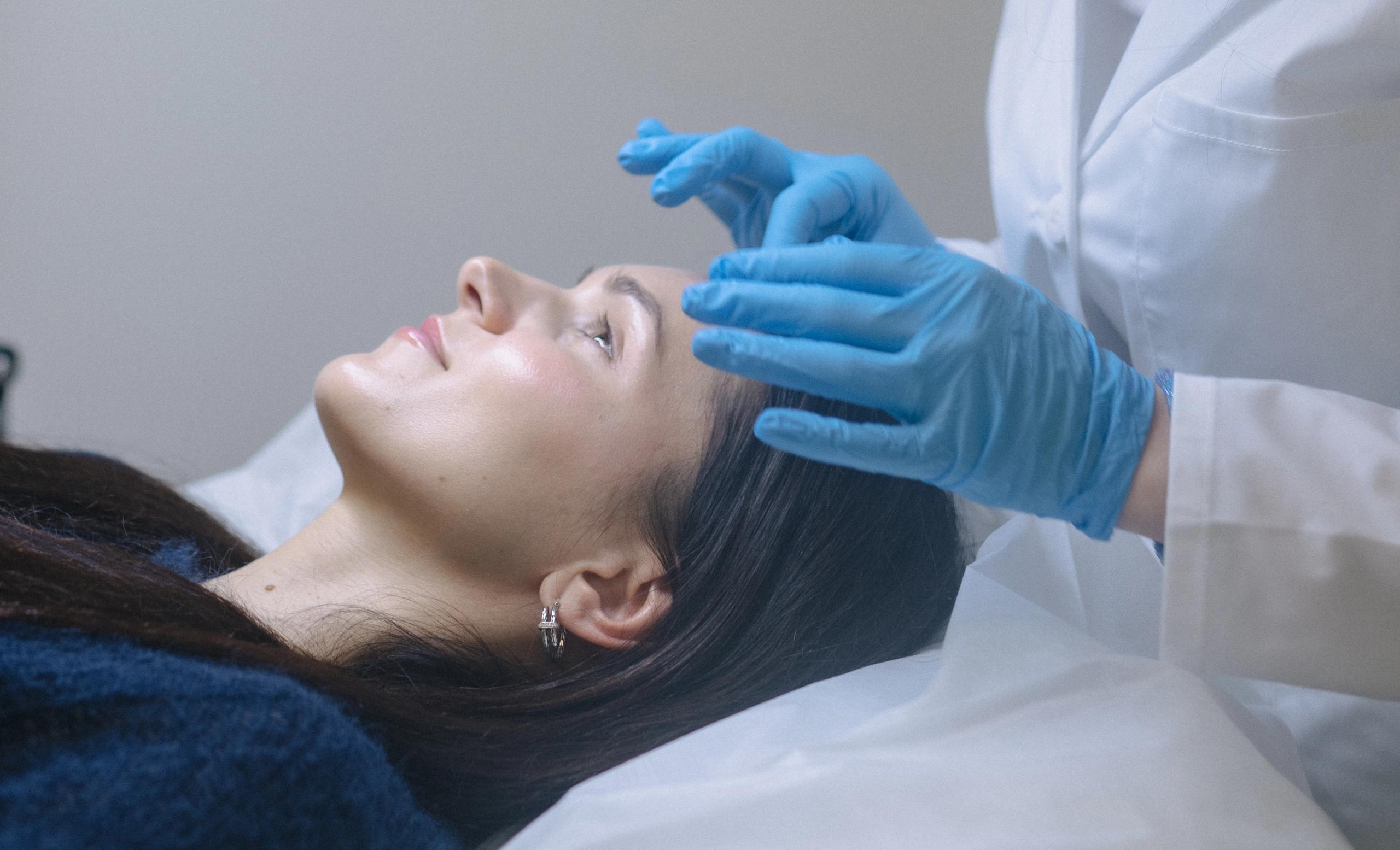 A person in blue gloves and a white lab coat gently touches a woman's forehead.