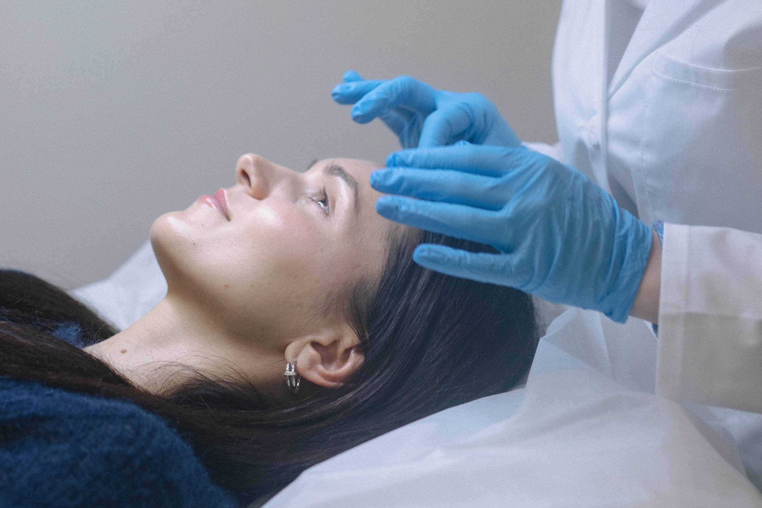 A person in blue gloves and a white lab coat gently touches a woman's forehead.