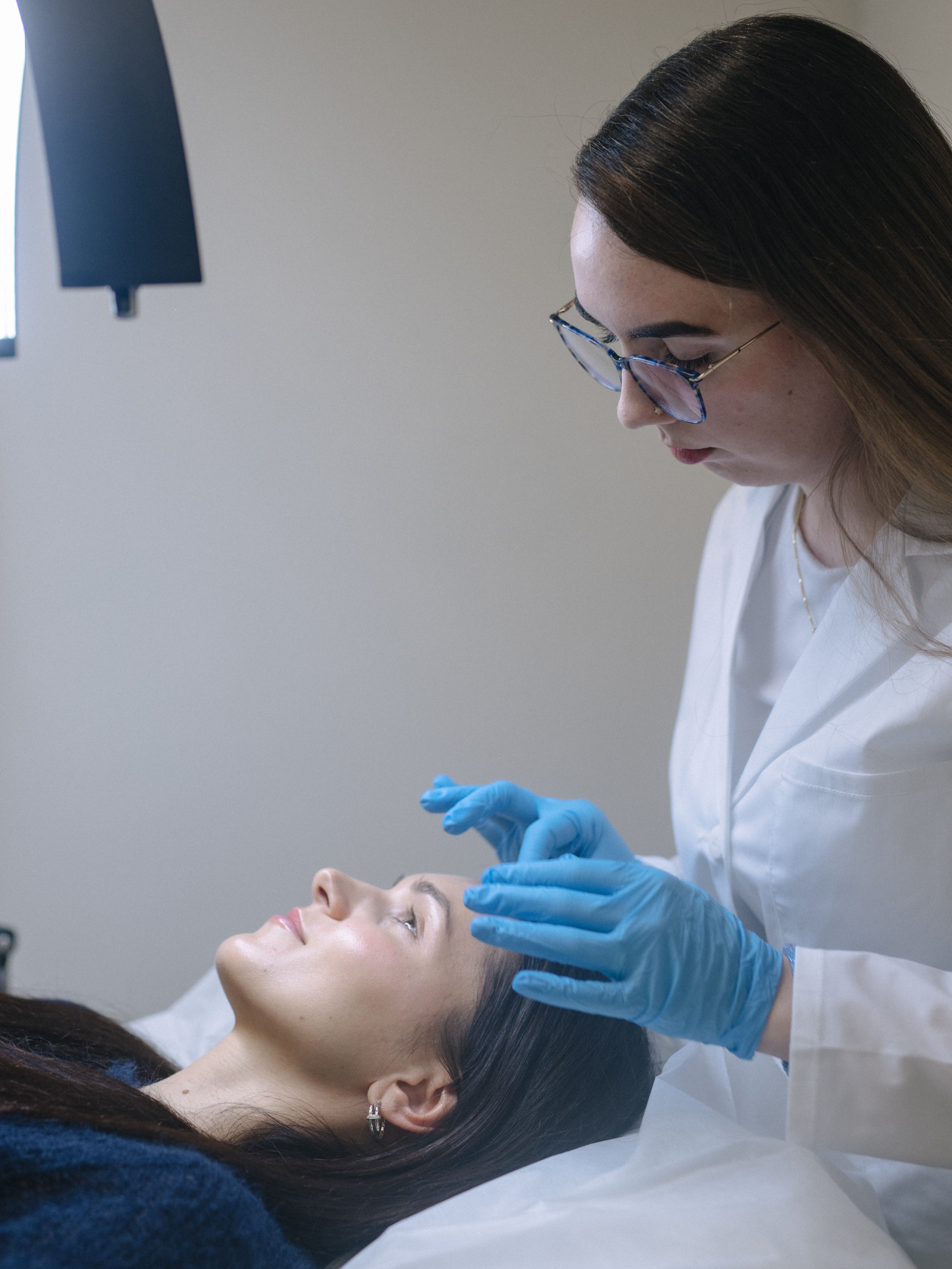 A practitioner in a lab coat and blue gloves examines a client's eyebrows
