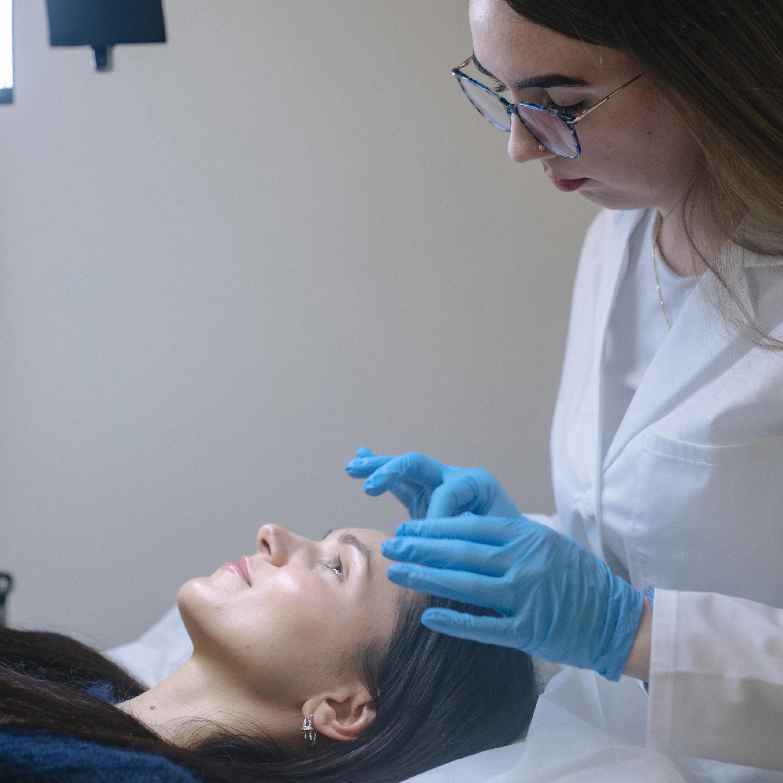 A practitioner in a lab coat and blue gloves examines a client's eyebrows