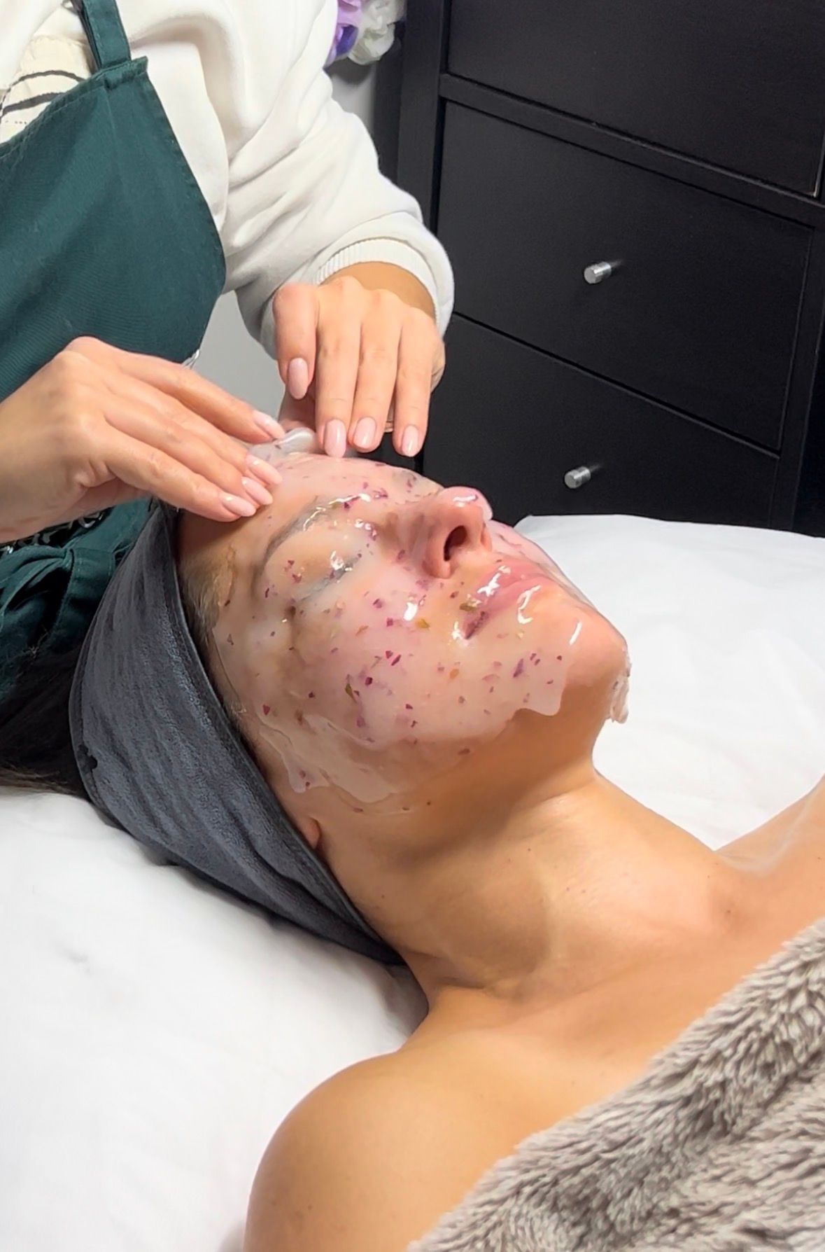 Hands applying a clear jelly mask with embedded flower petals to a person's face.