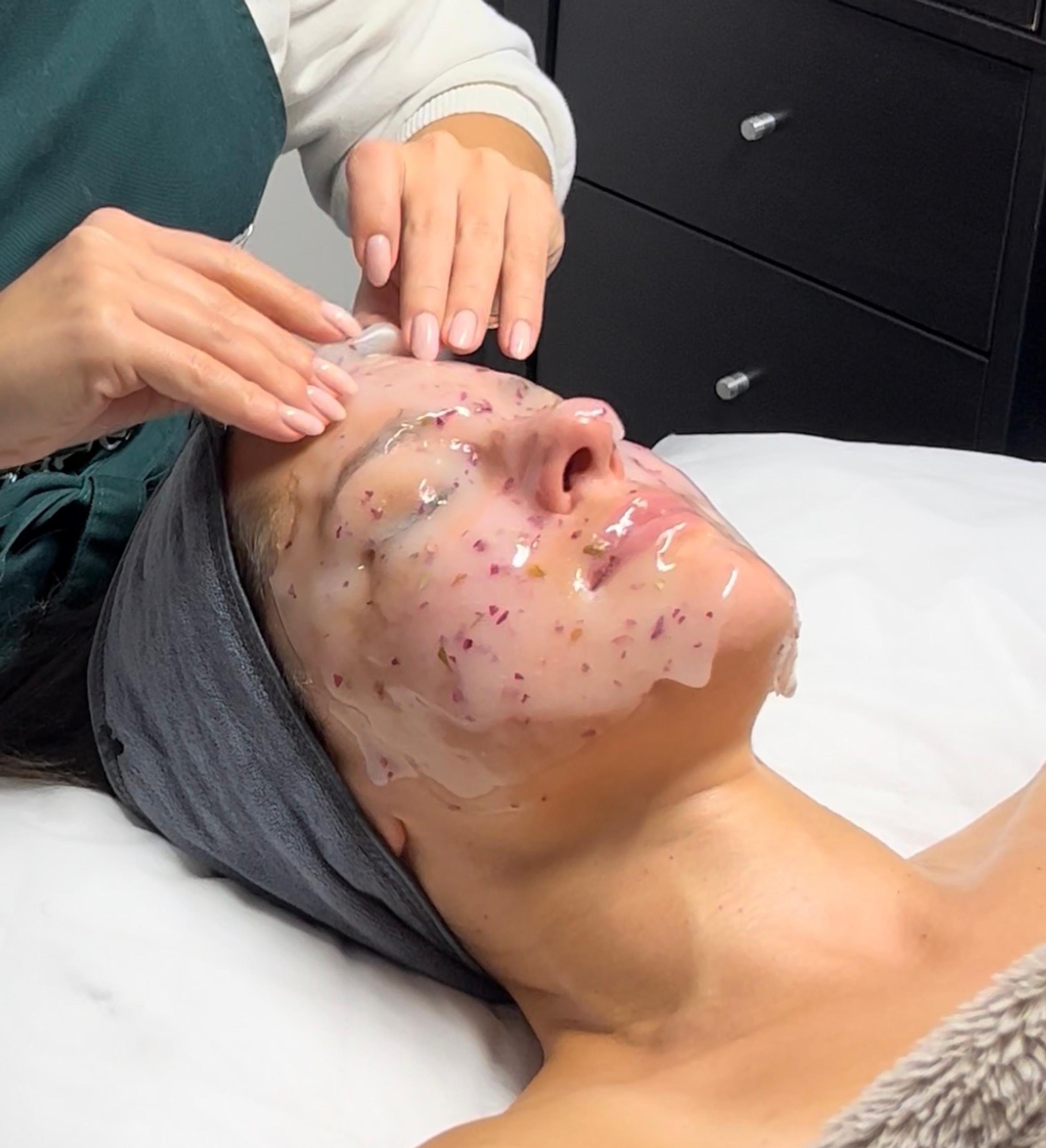 Hands applying a clear jelly mask with embedded flower petals to a person's face.