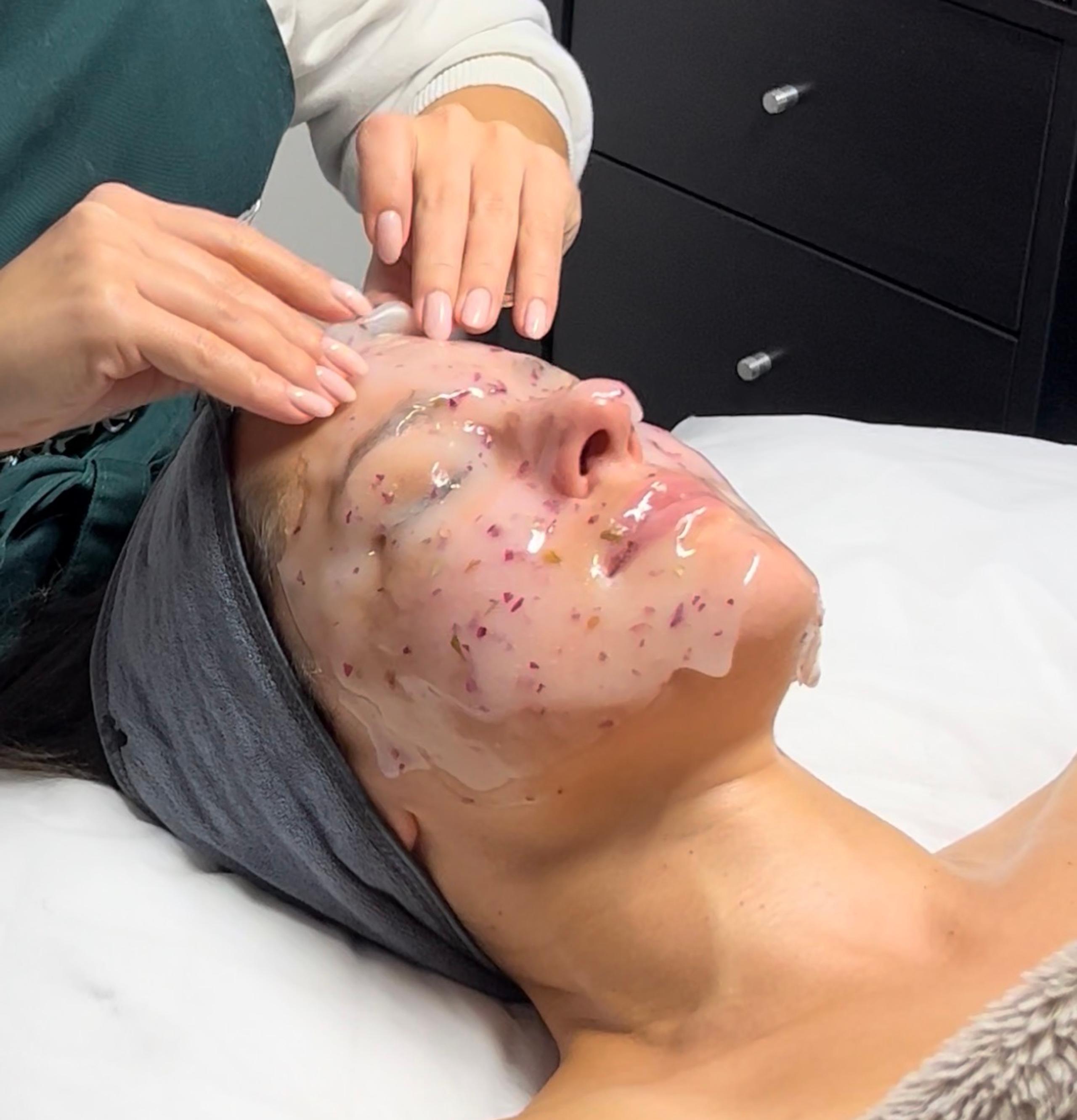 Hands applying a clear jelly mask with embedded flower petals to a person's face.