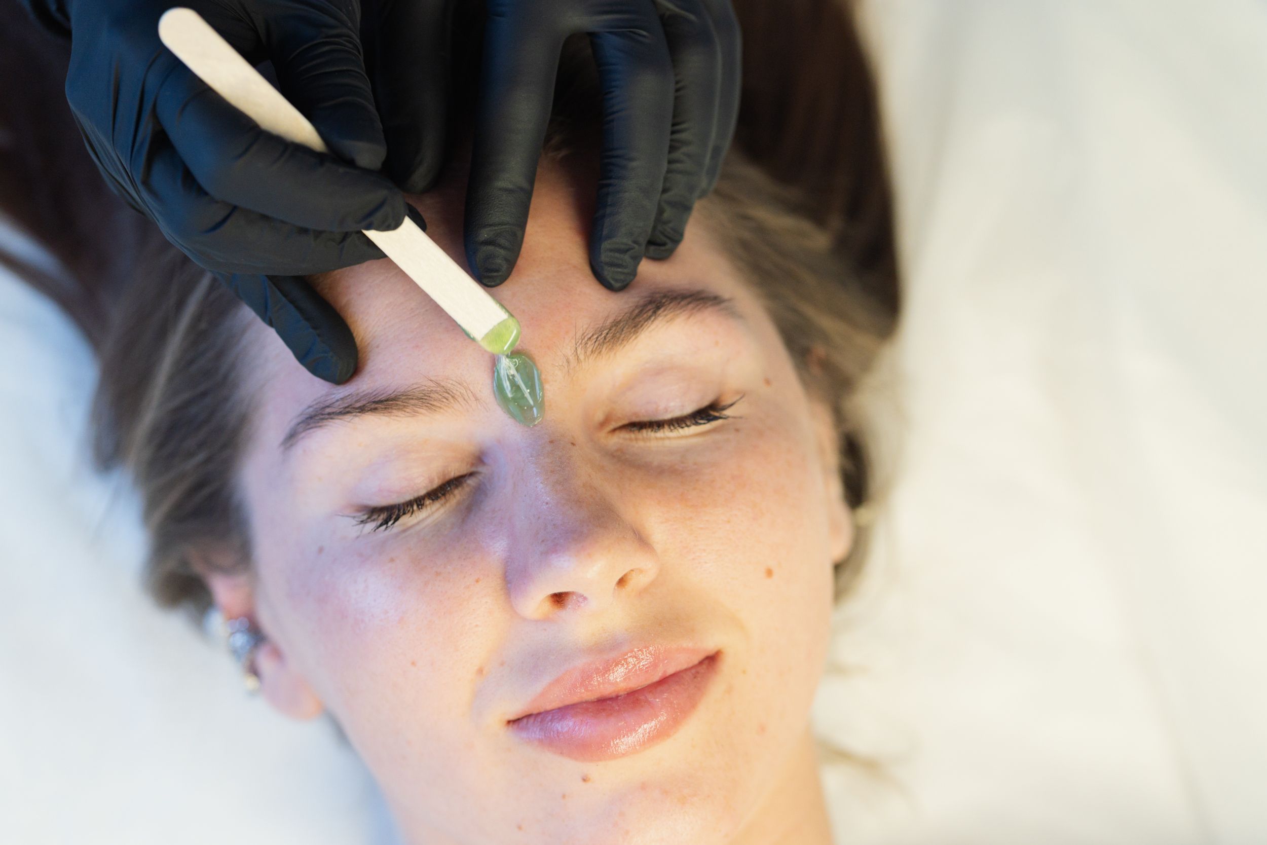 A gloved hand applies green wax to a woman's forehead for eyebrow waxing.