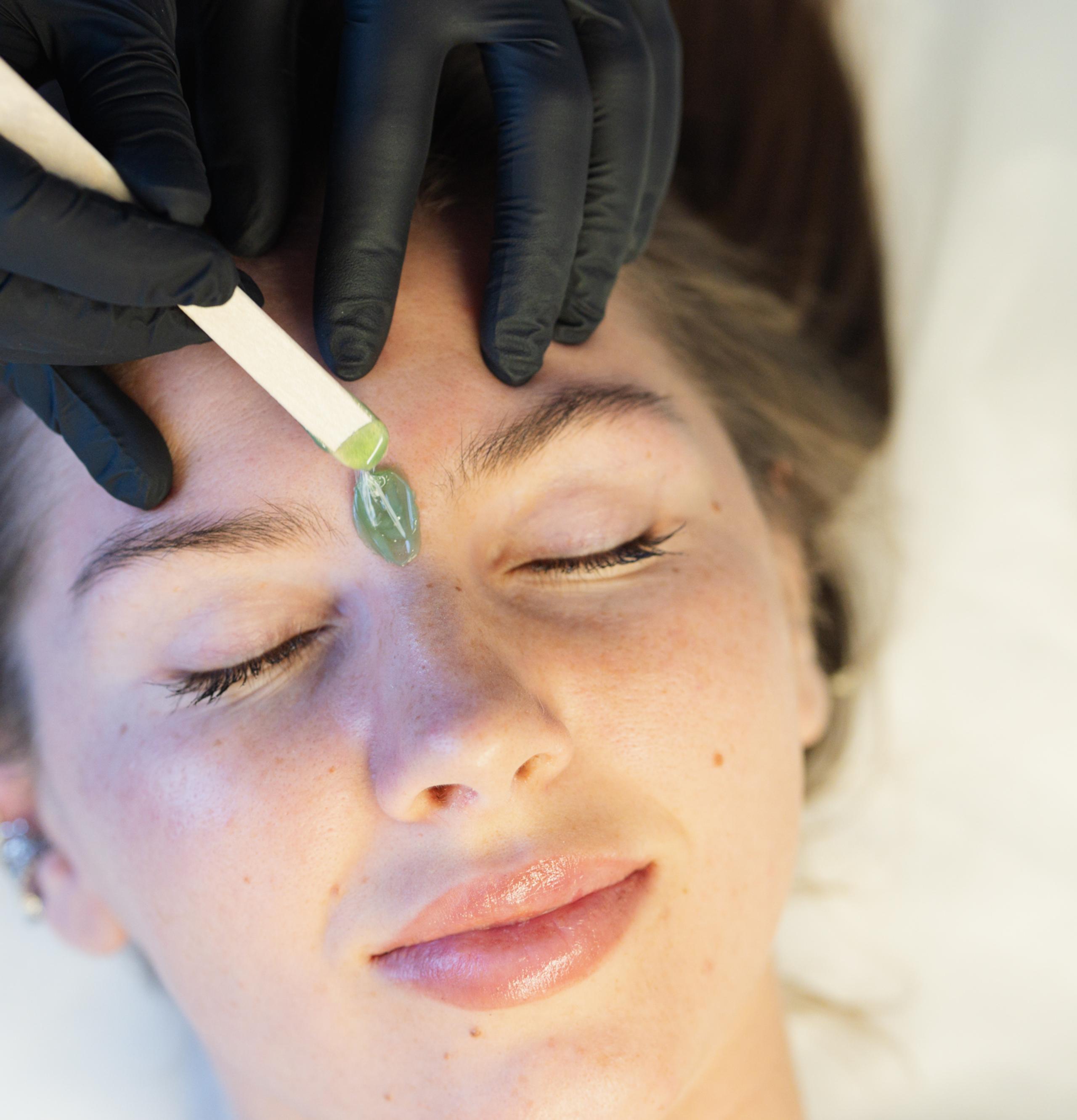 A gloved hand applies green wax with a stick to a woman's forehead.