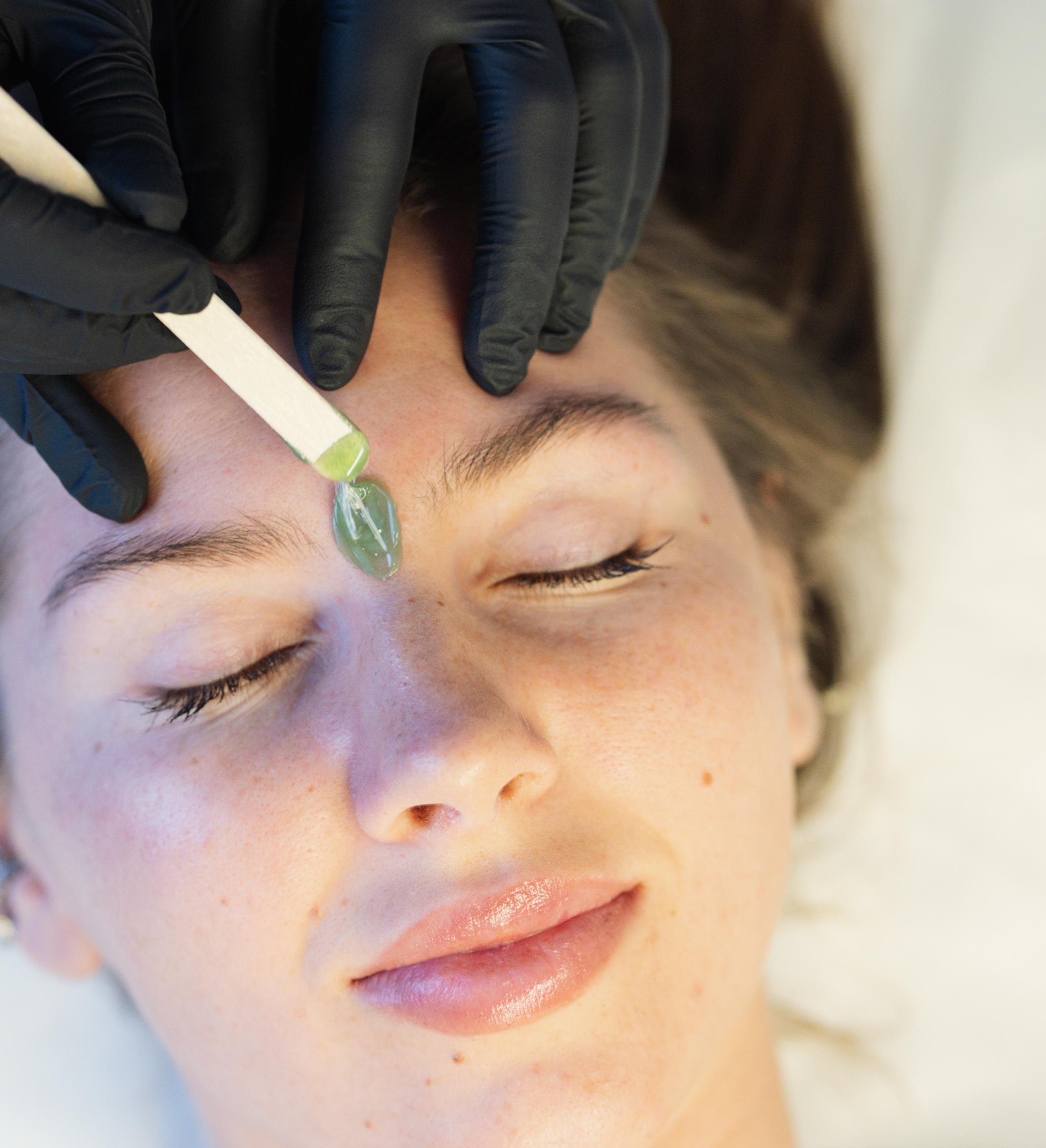 Gloved esthetician applying green wax between a client's eyebrows with a wooden spatula.