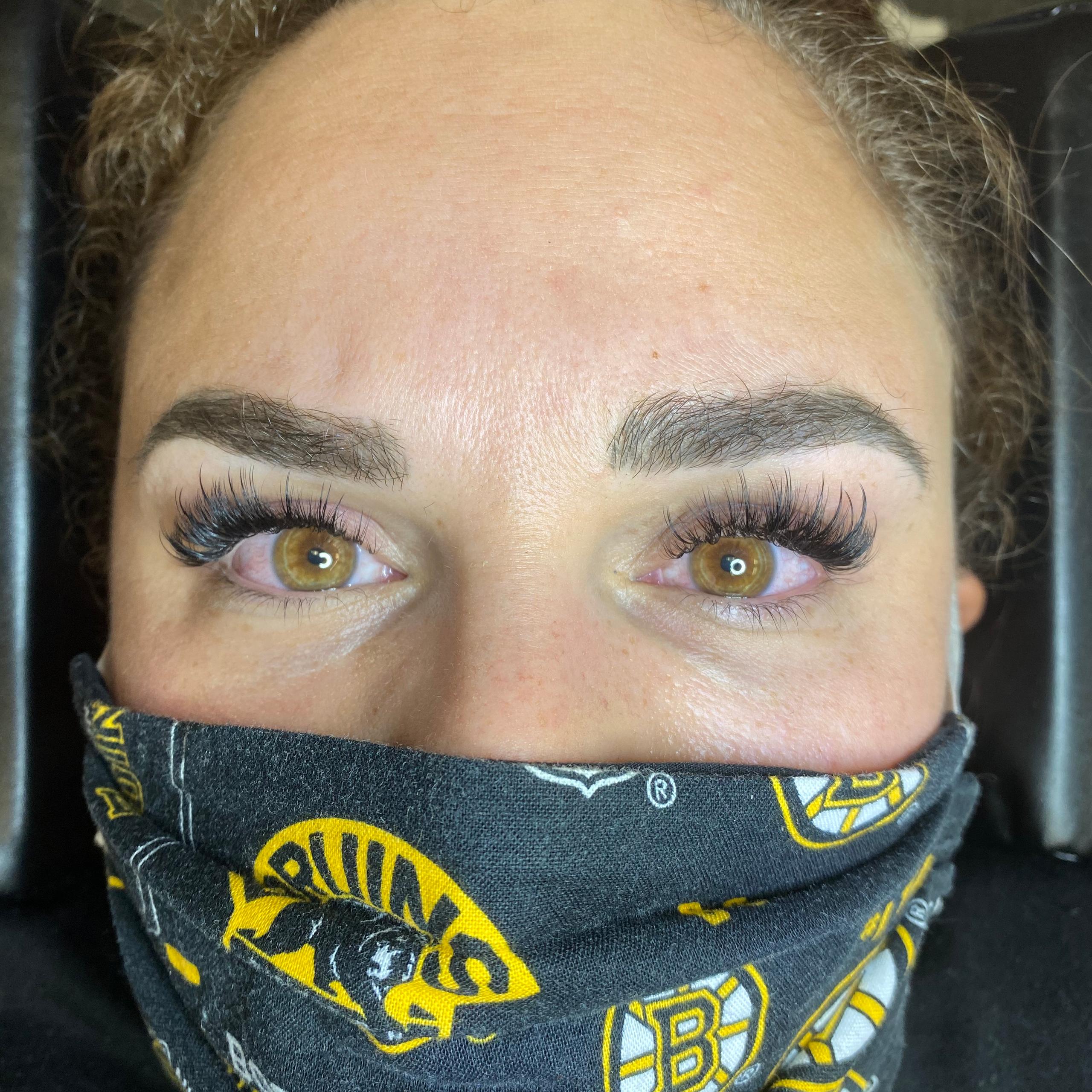 A person's face with full, dark eyelashes, defined eyebrows, and wearing a black face mask.