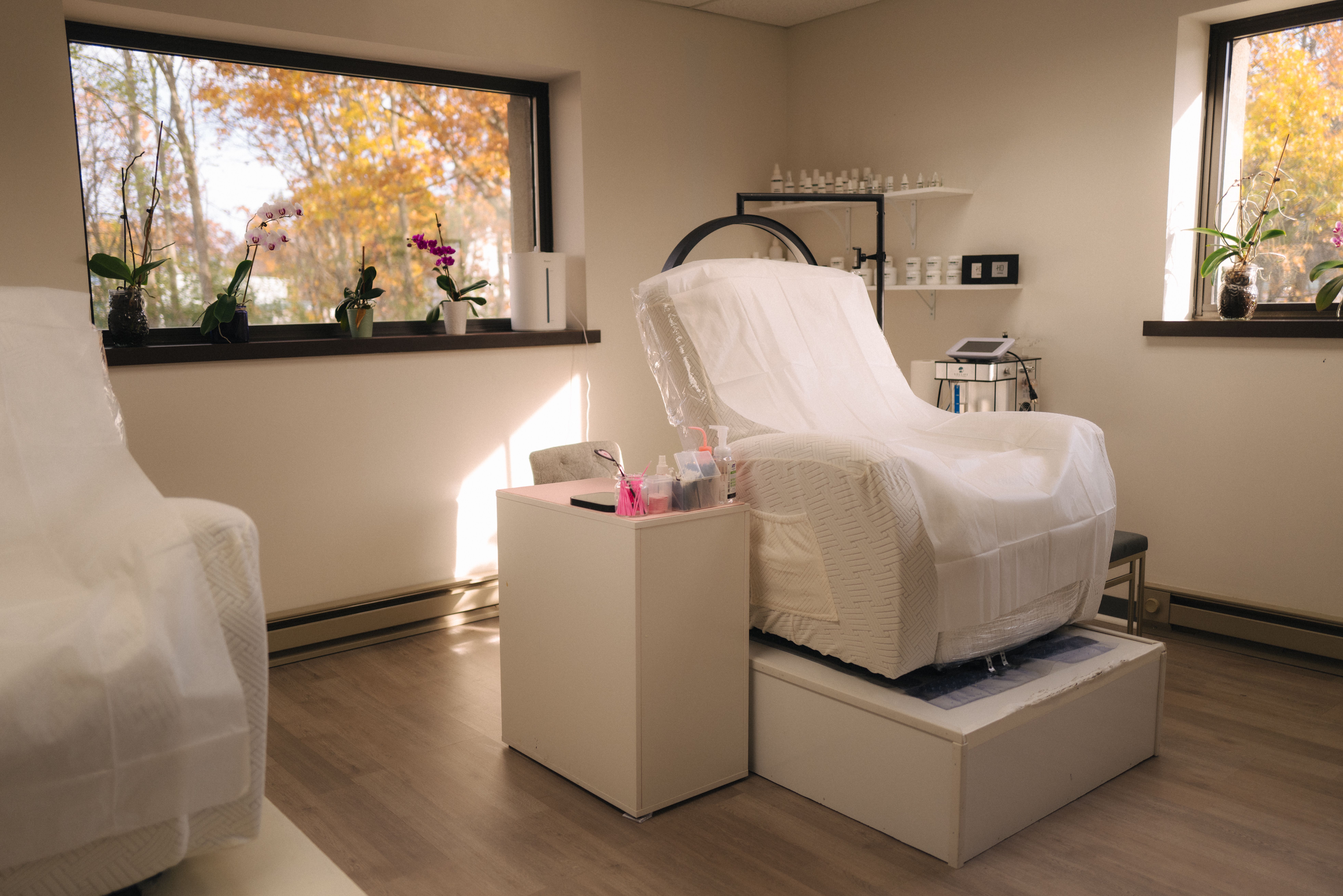 A clean, brightly lit treatment room featuring a white covered treatment chair on a raised platform, with autumn trees visible through two windows.