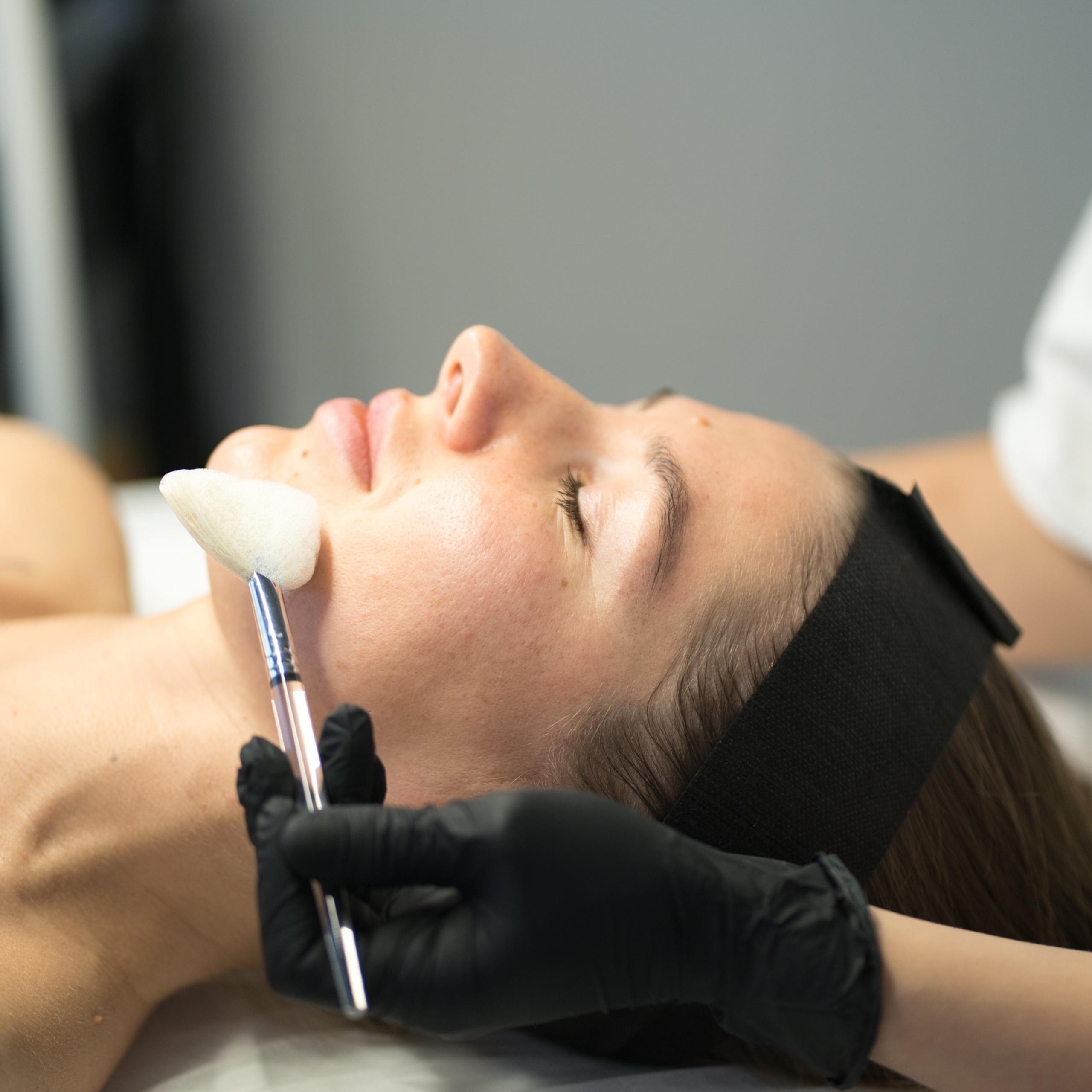 A gloved hand applies a facial product to a woman's cheek with a brush.