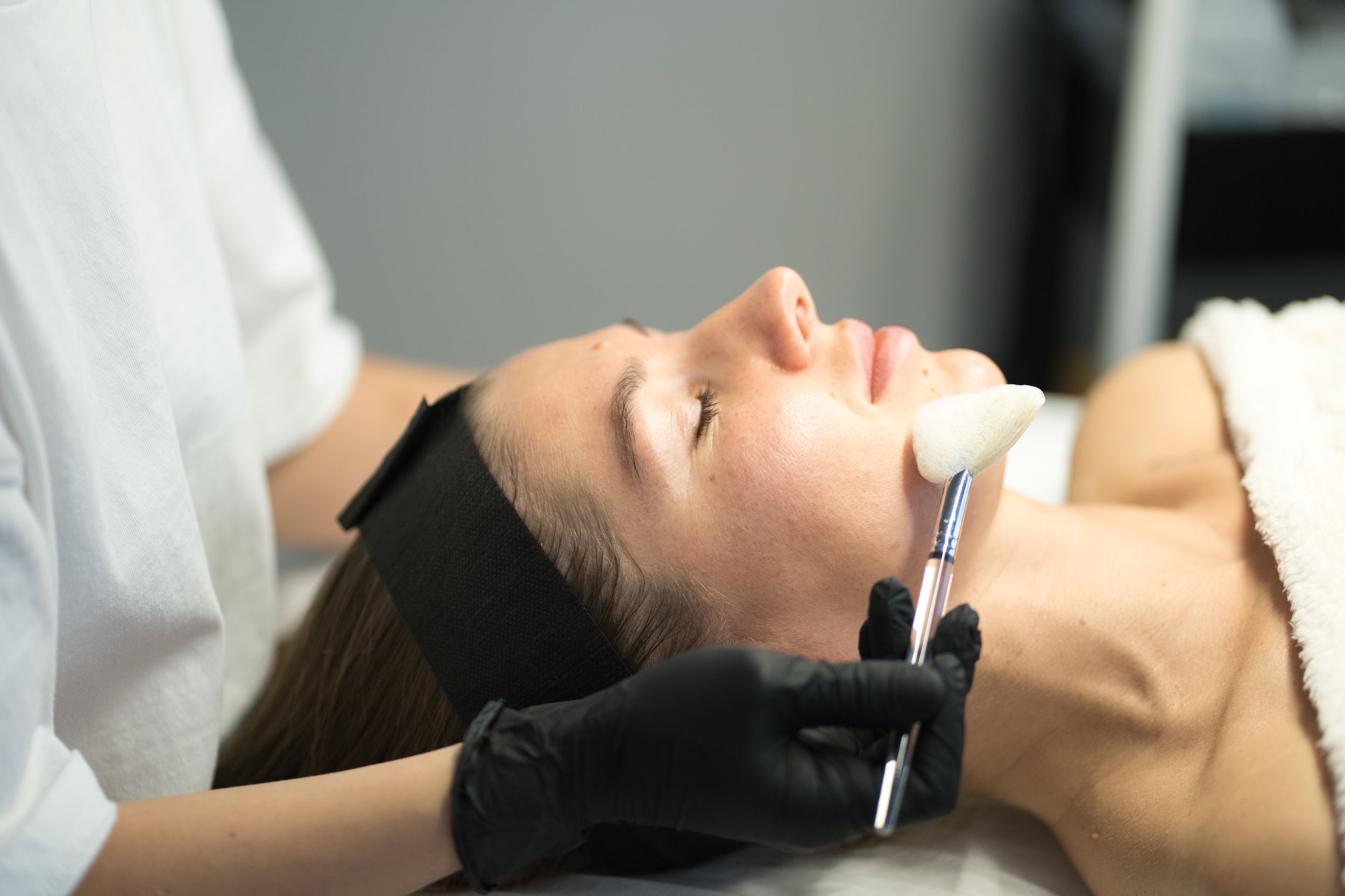 A woman lies with eyes closed as an aesthetician applies a facial product to her cheek with a brush.