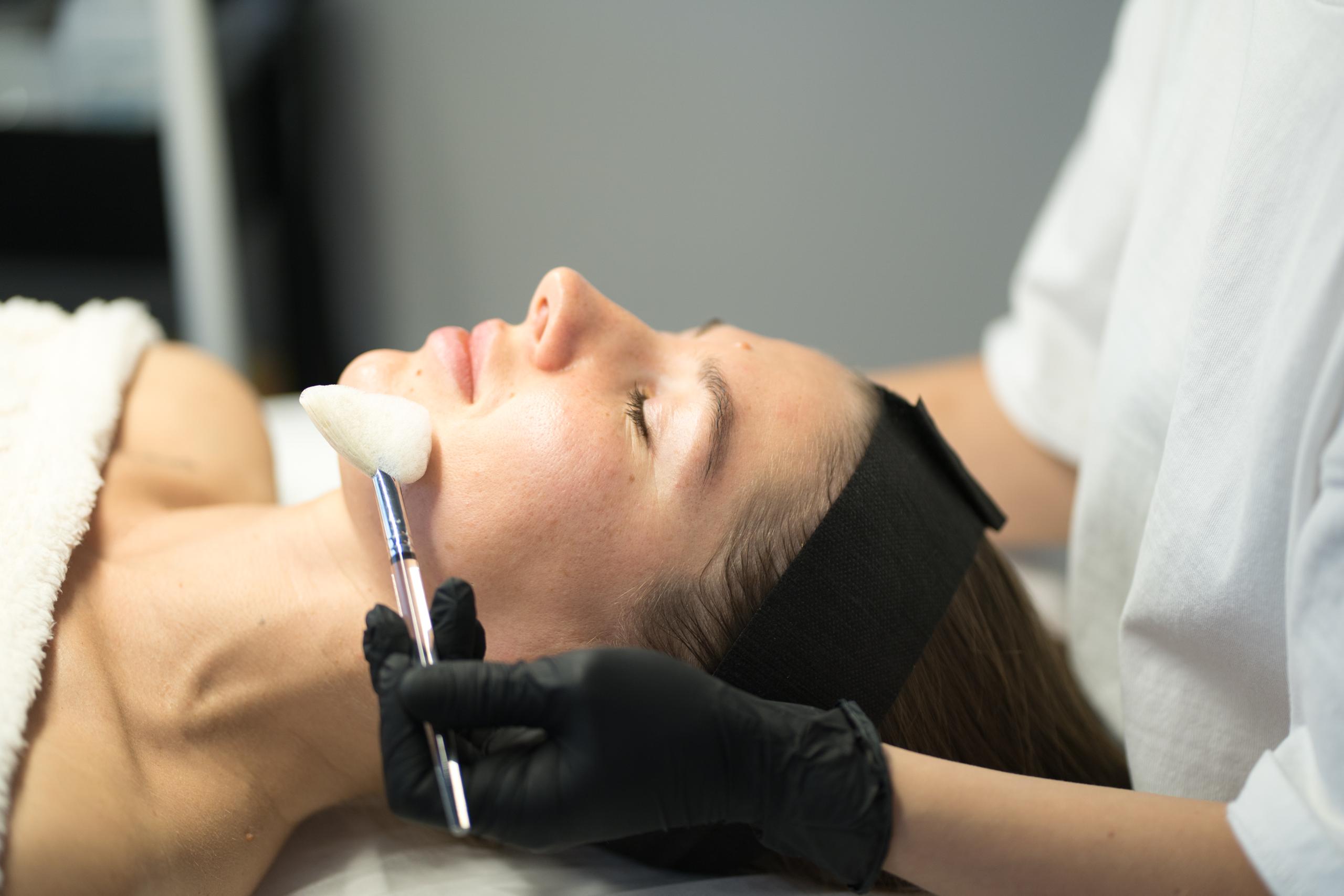 A woman lies with eyes closed as an aesthetician applies a facial product to her cheek with a brush.