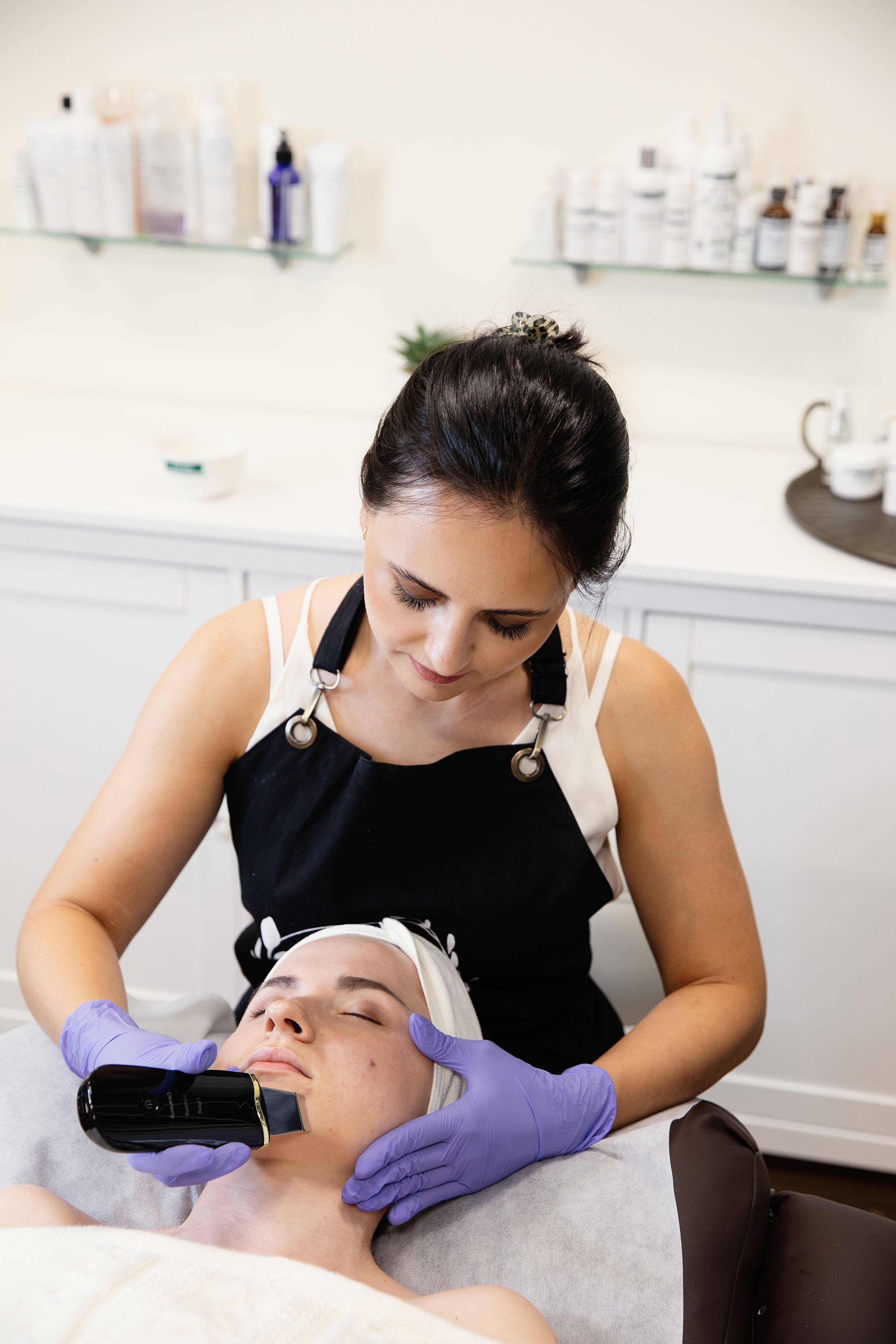 An esthetician in purple gloves uses a black skin scrubber on a client's face. 
