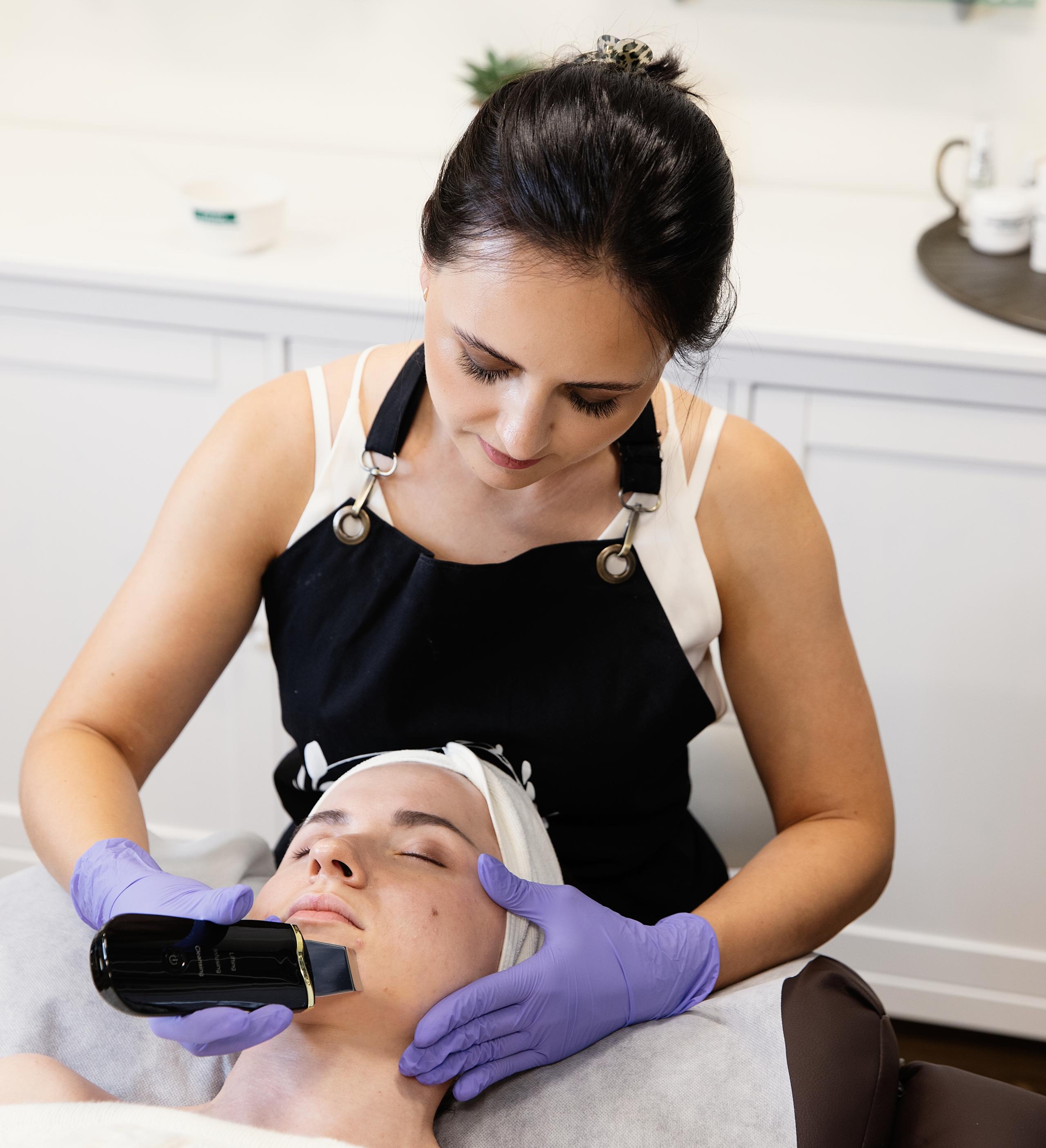 An esthetician in purple gloves uses a black skin scrubber on a client's face.